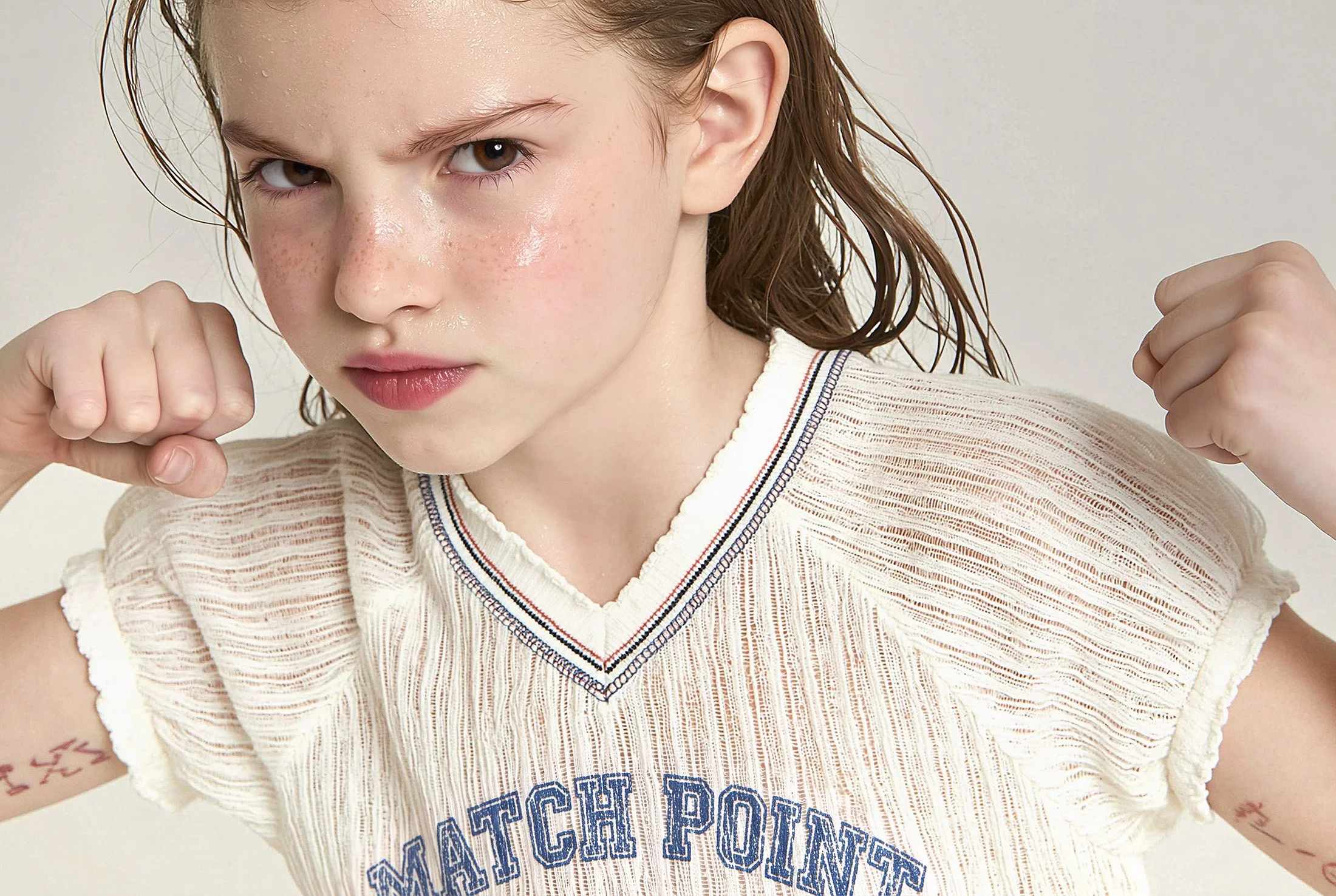 Girl in a cream textured knit tennis vest with navy and red tipping, wet hair against a grey seamless — kidswear editorial, varsity sports print, close-up, AI-generated image.