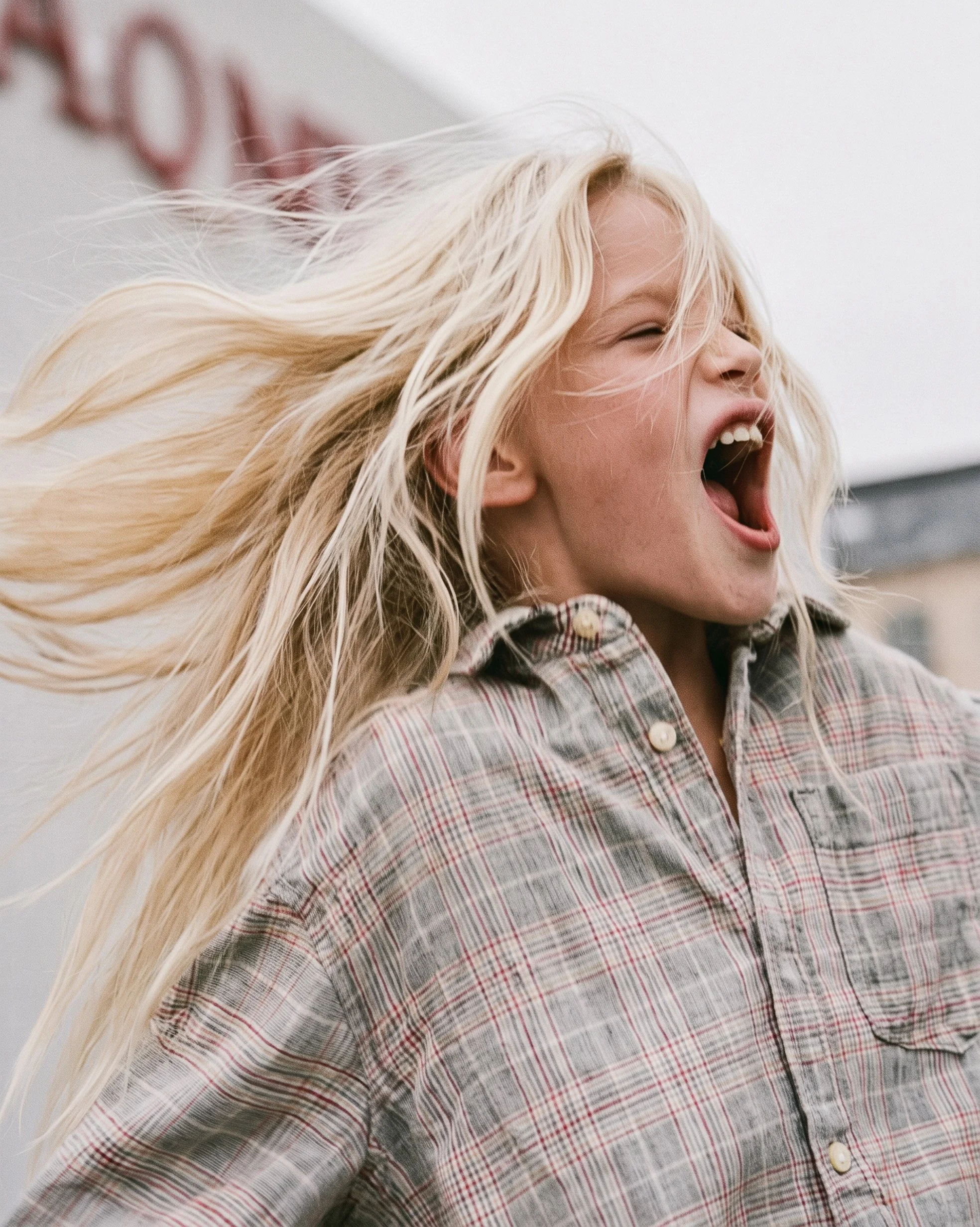 Girl shouting with her eyes shut and hair flying, grey and red checked cotton shirt with pearl snap buttons — kidswear lifestyle editorial, overcast daylight. AI-generated image.