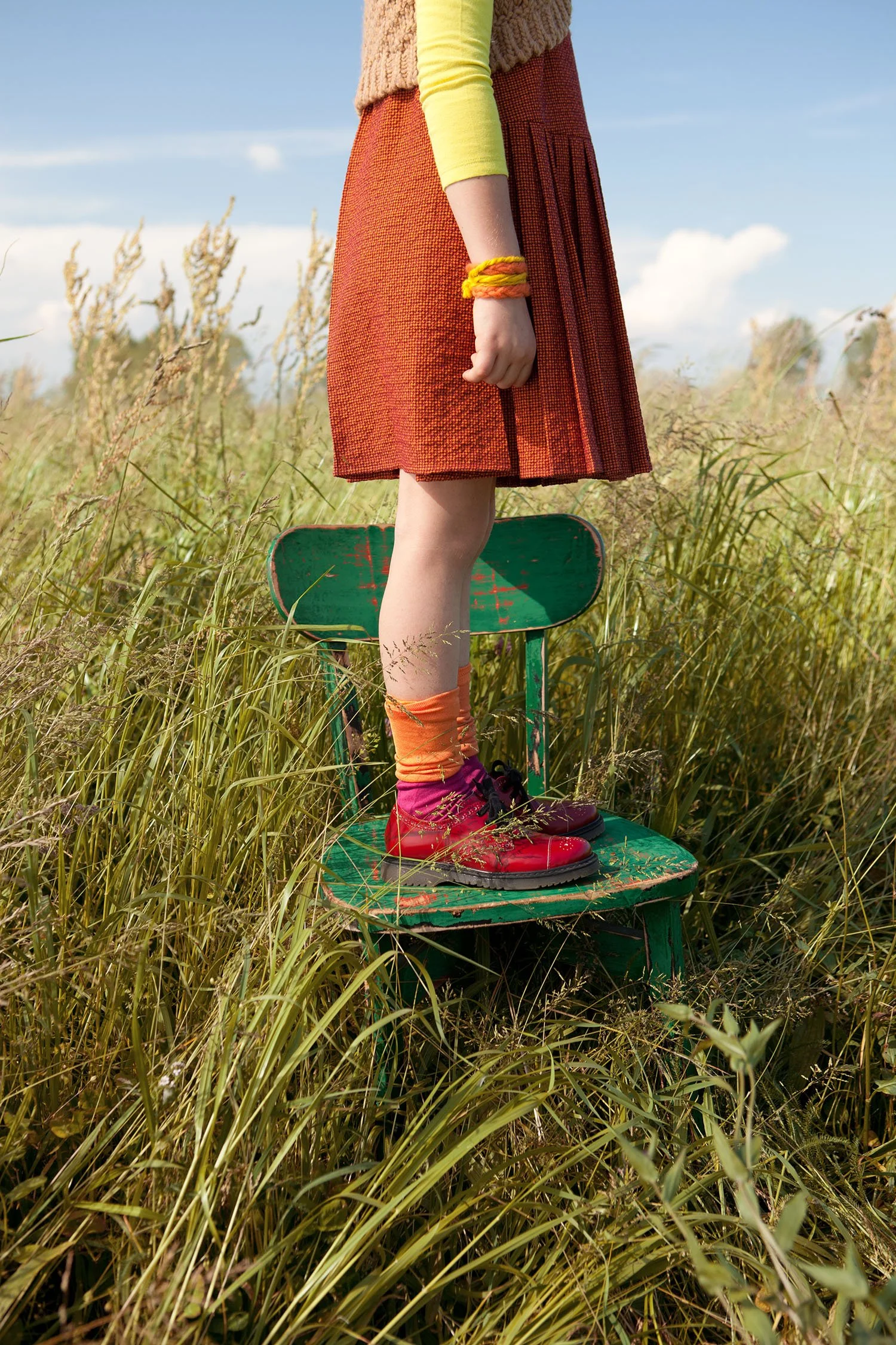 Style Piccoli editorial — orange pleated skirt on a green stool in tall grass.