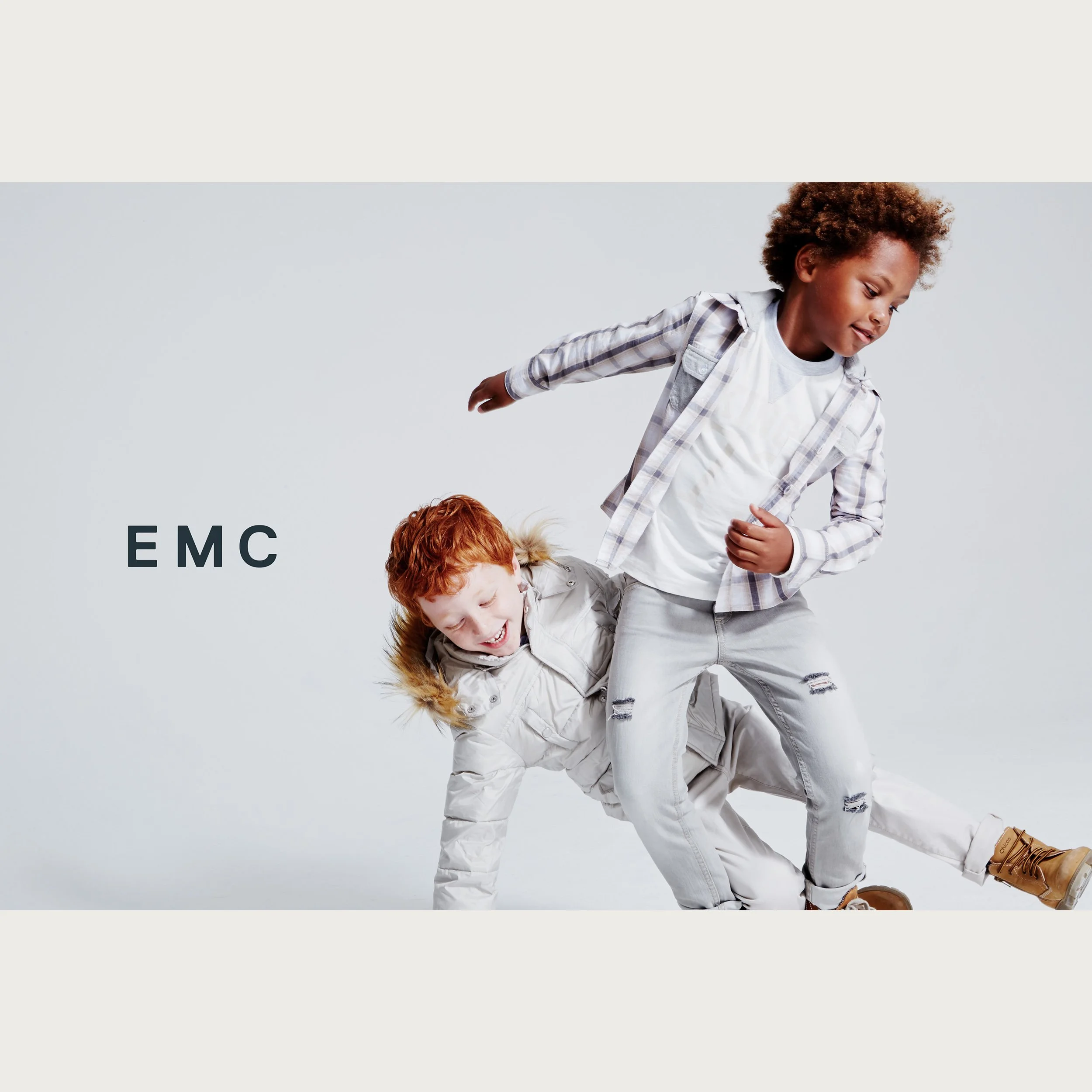 Ivory down jacket with checked shirt, two boys play-jumping on pale grey — EMC campaign.
