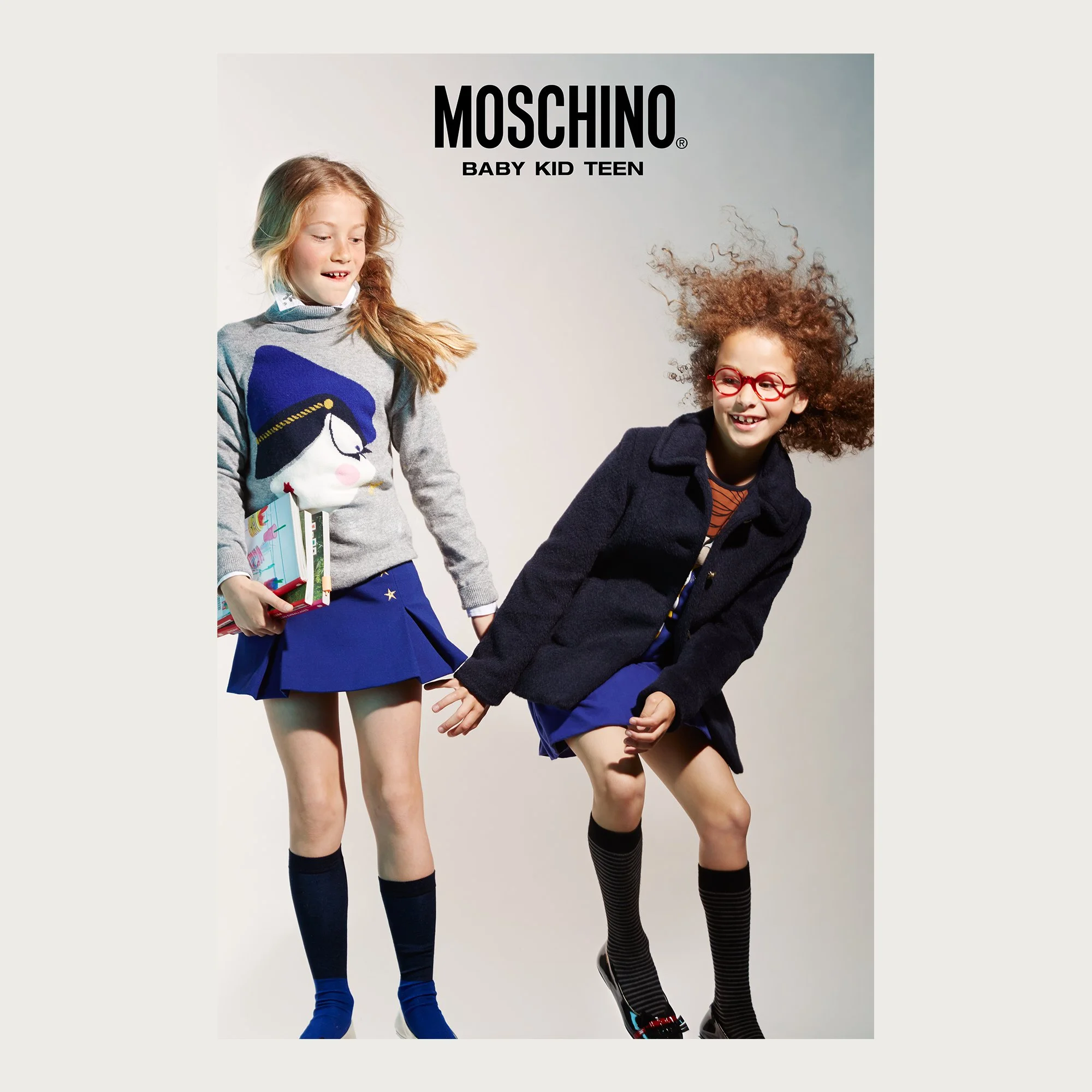 Cobalt pleated skirts, navy coat and intarsia knit, two girls on a grey backdrop — Moschino Kid campaign.