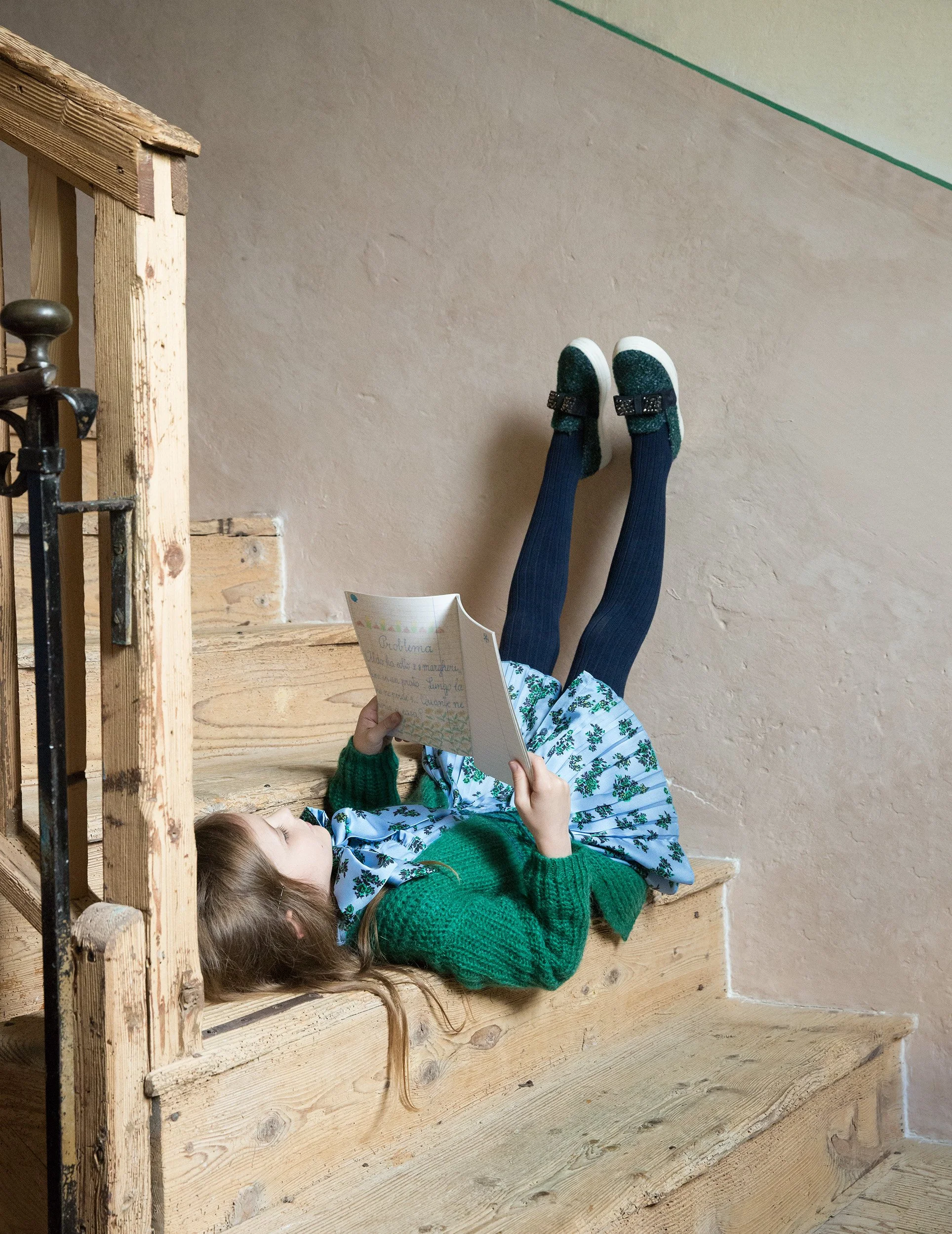 Style Piccoli editorial — child reading upside down on a wooden stair, feet against the wall.