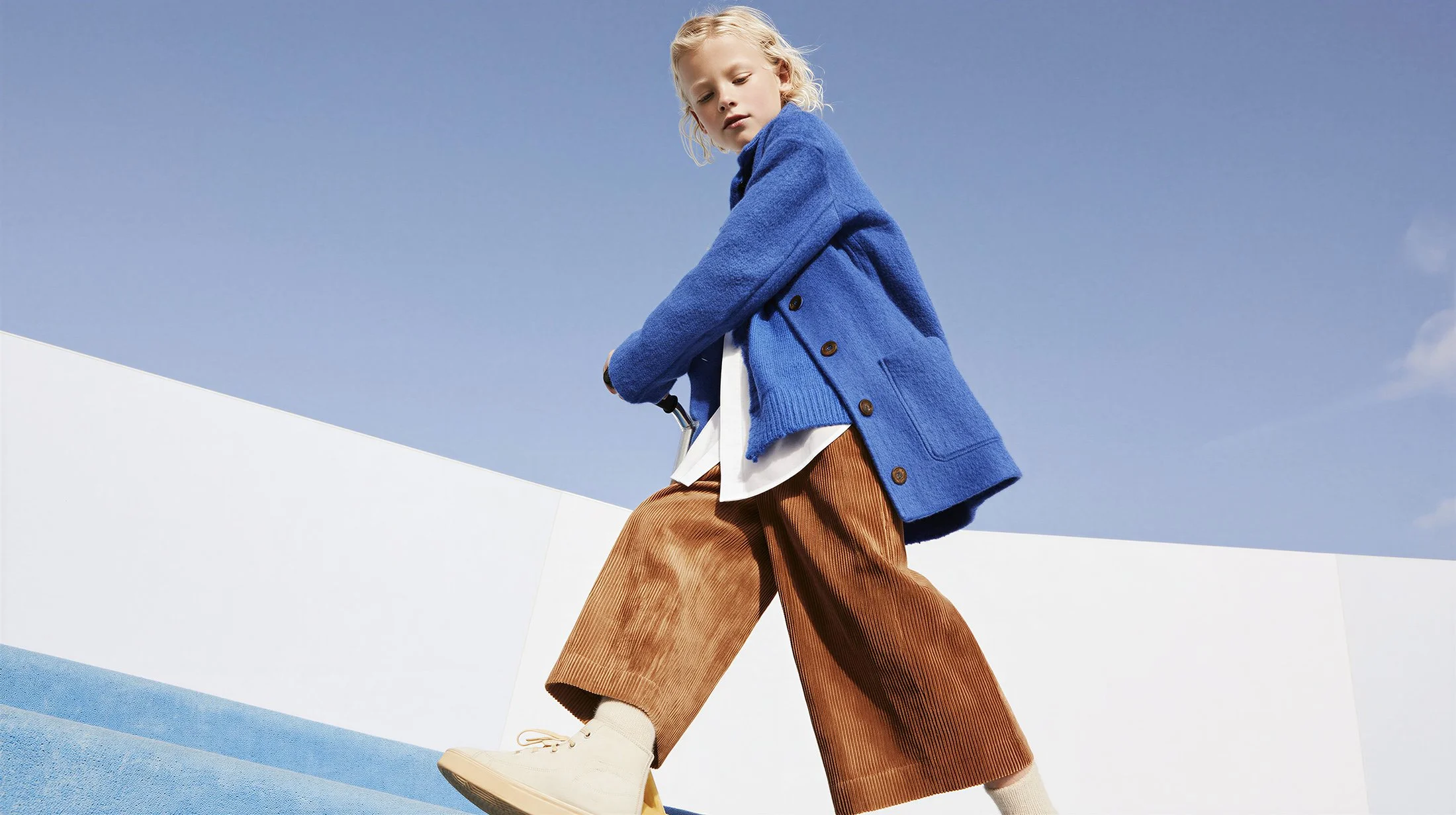 Child walking in an oversized blue wool coat and camel trousers. AI-generated image.