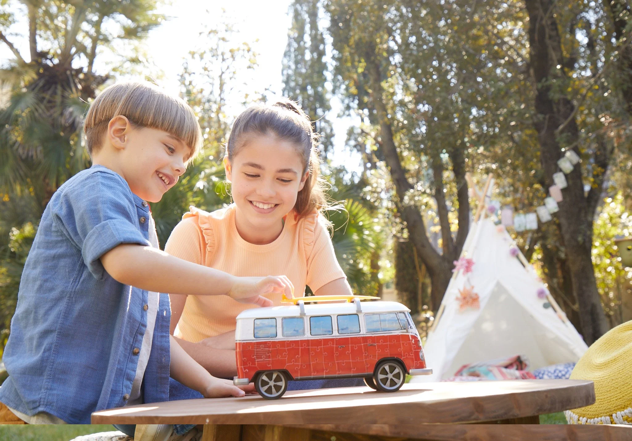 Two children assembling a Ravensburger 3D campervan puzzle on a garden table, teepee and bunting behind — toy advertising, outdoor lifestyle, warm backlight.