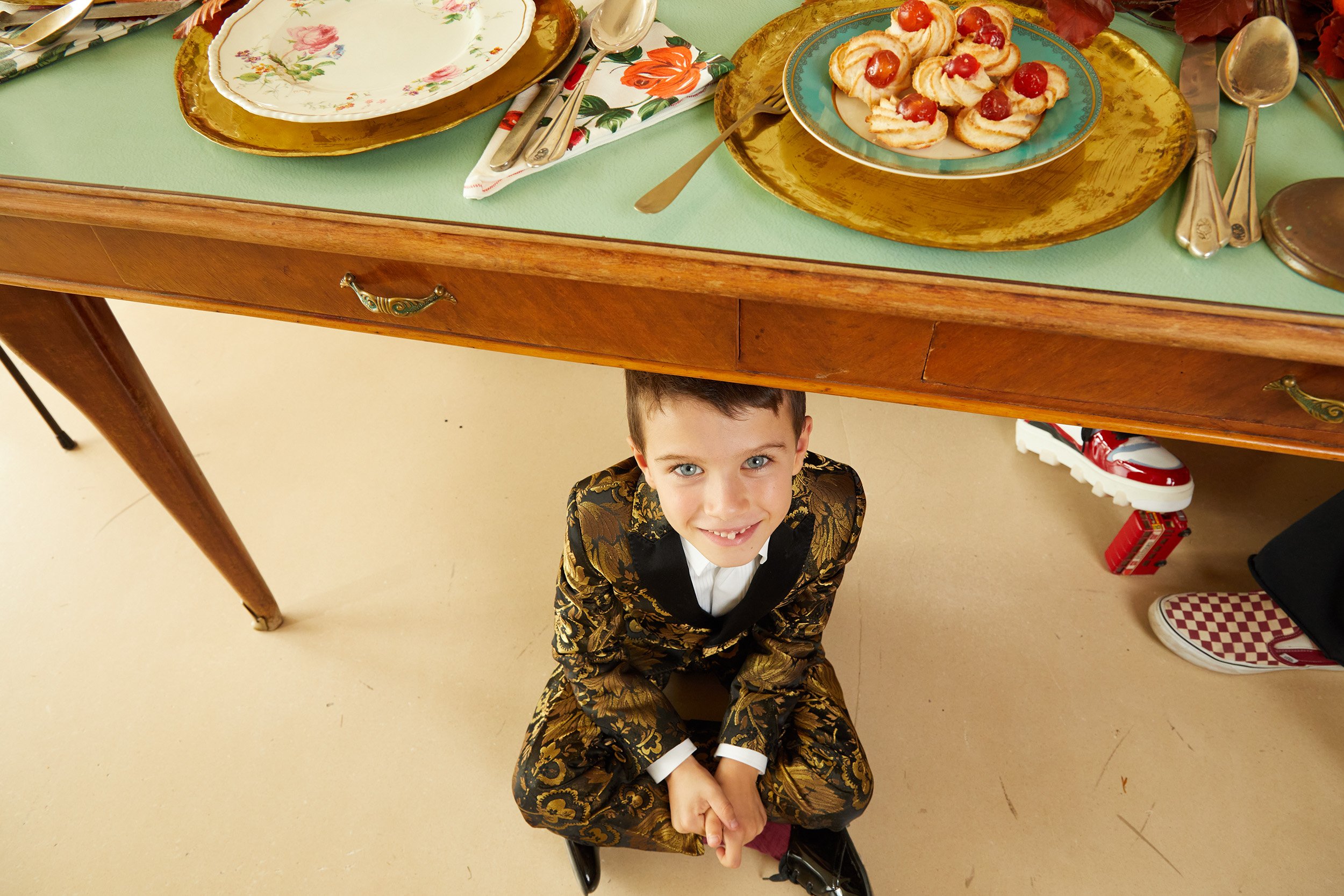 Boy in a brocade jacket sitting under a laid vintage table.