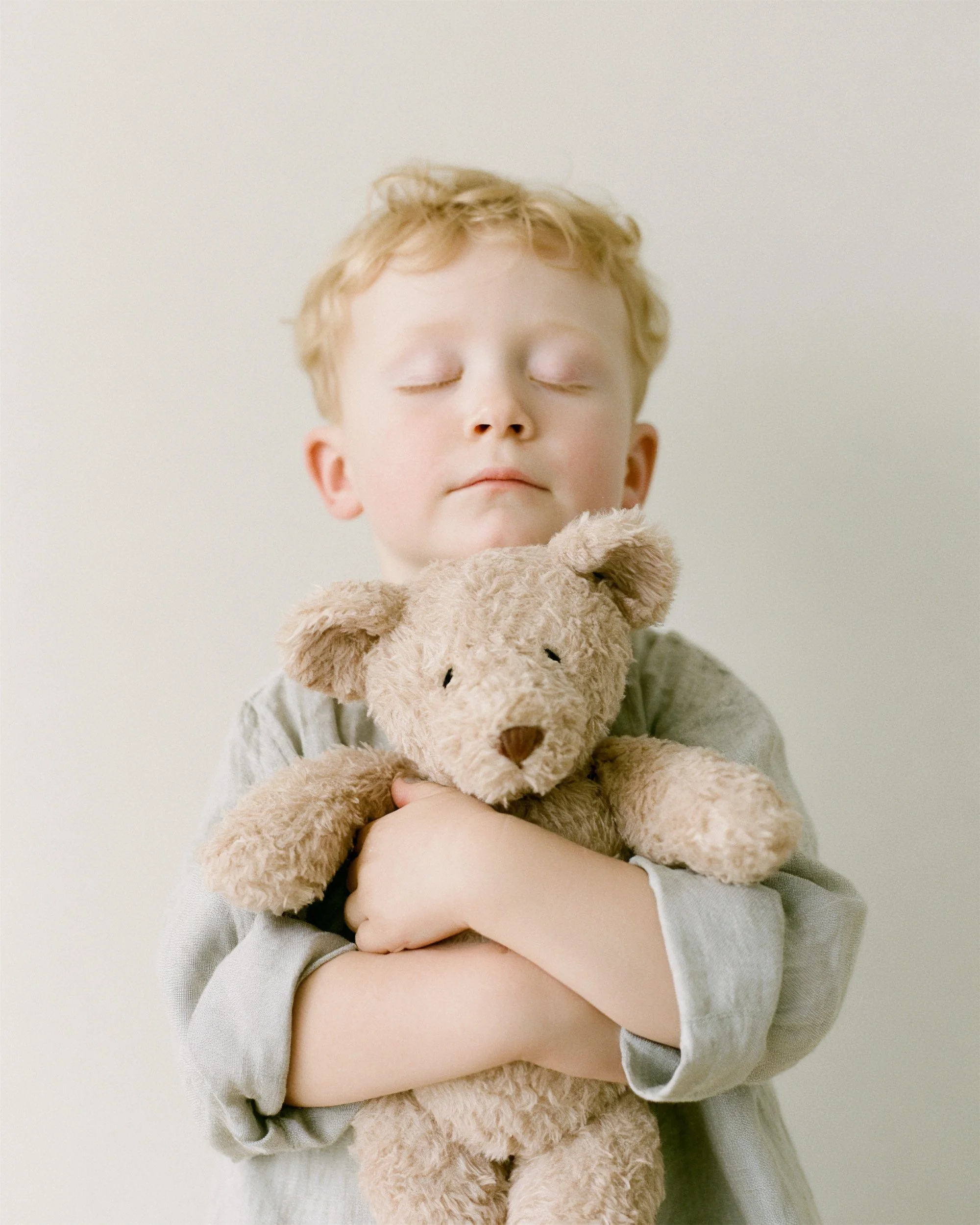 Baby with eyes closed hugging a teddy bear, soft neutral tones. AI-generated image.