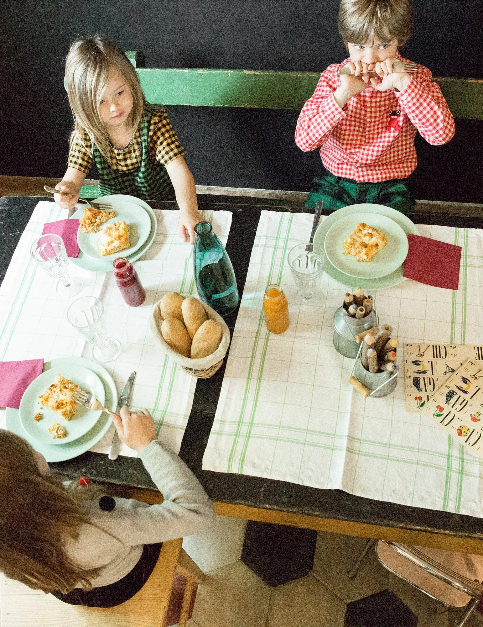 Style Piccoli editorial — two children eating at a laid table, seen from overhead.