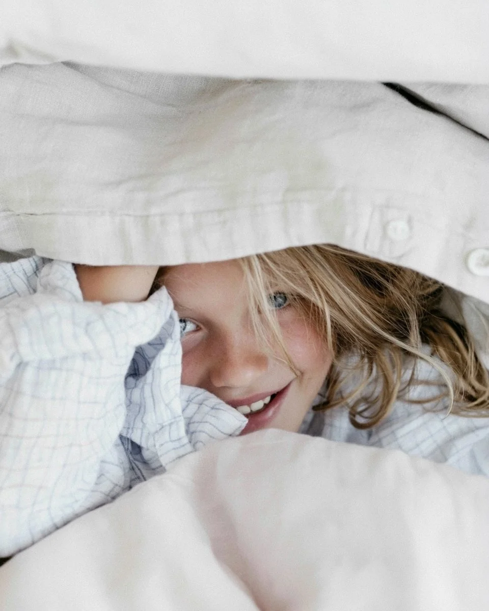 Child peeking out from under a linen duvet in checked cotton pyjamas, white and natural bed linen — kidswear sleepwear and bed linen, lifestyle, AI-generated image.