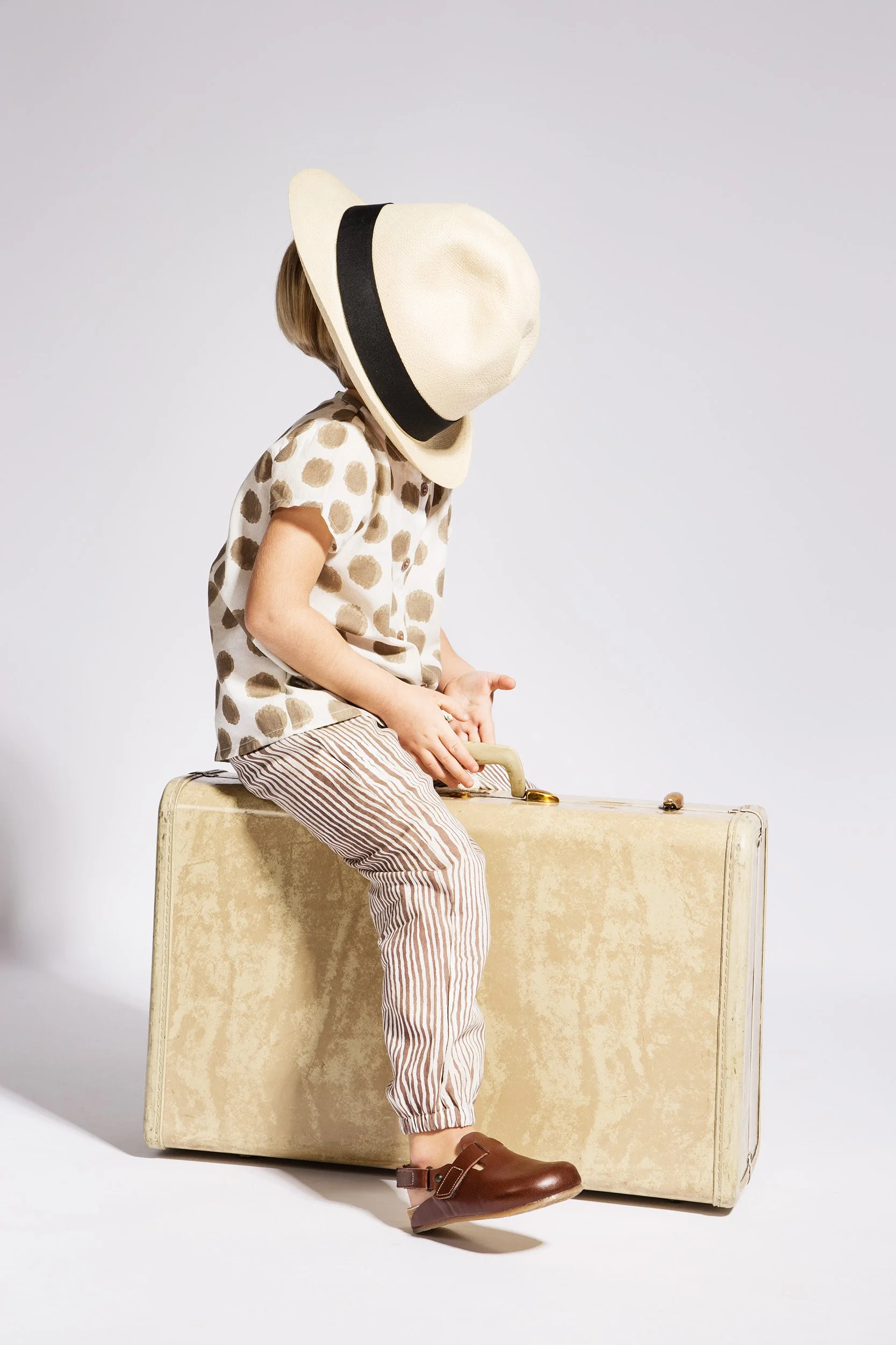Child in a straw hat sitting on a vintage suitcase with a dot shirt, beige tones.