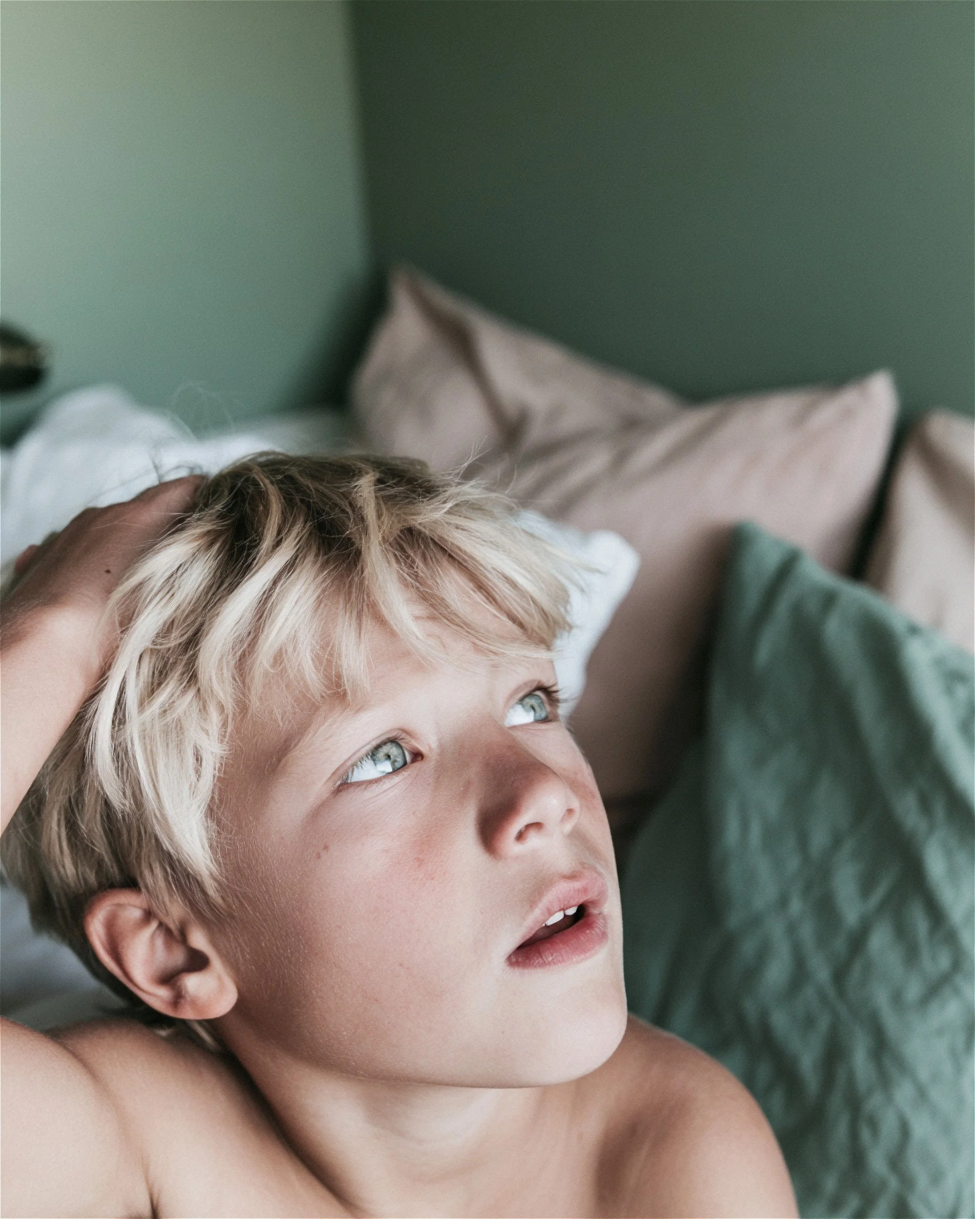 Boy on a bed looking up, sage green wall, dusty pink pillow and quilted bedspread — lifestyle interiors and bed linen, soft daylight. AI-generated image.
