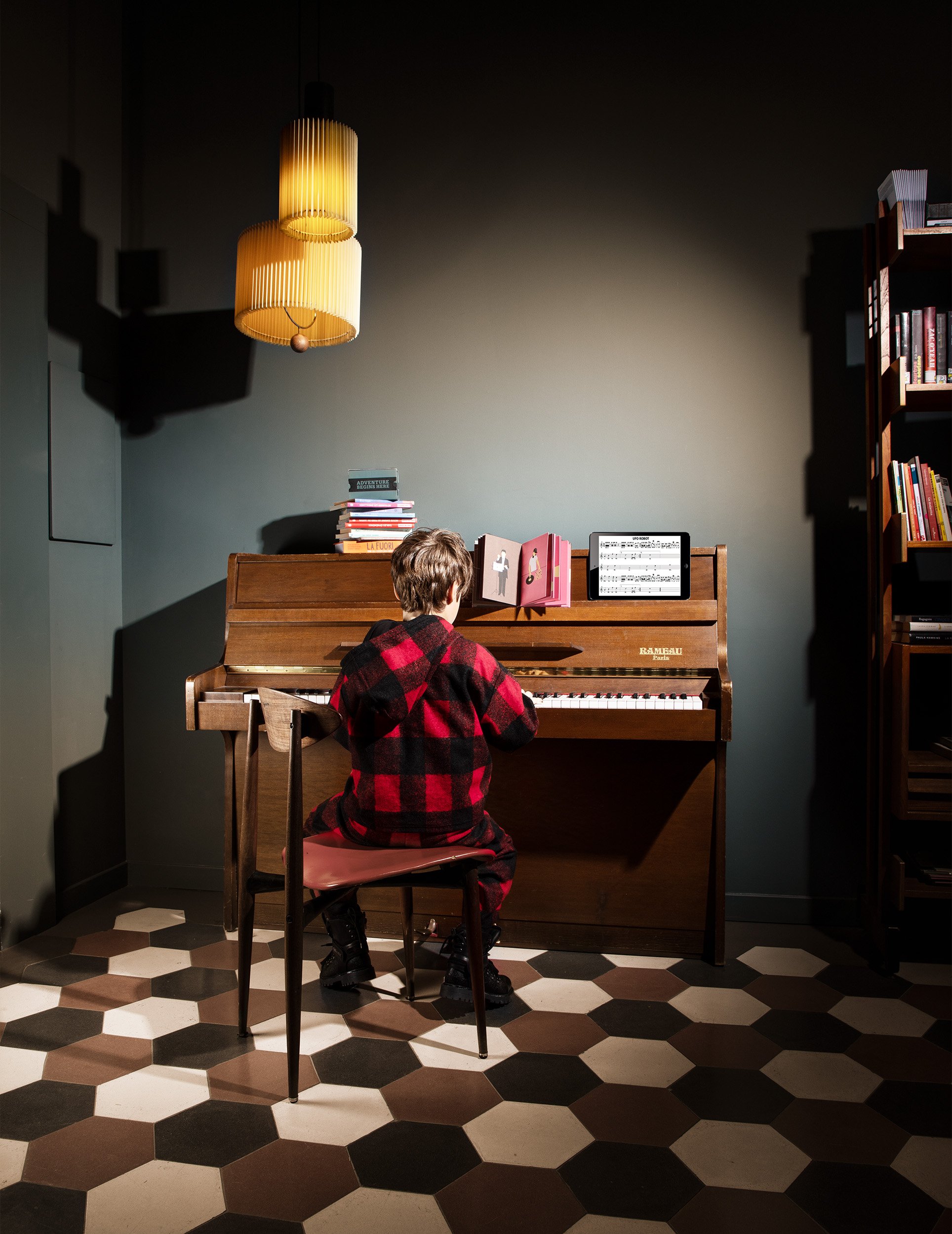 Child in red buffalo check at piano, teal walls and hexagonal cement tiles — kidswear, vintage interiors.