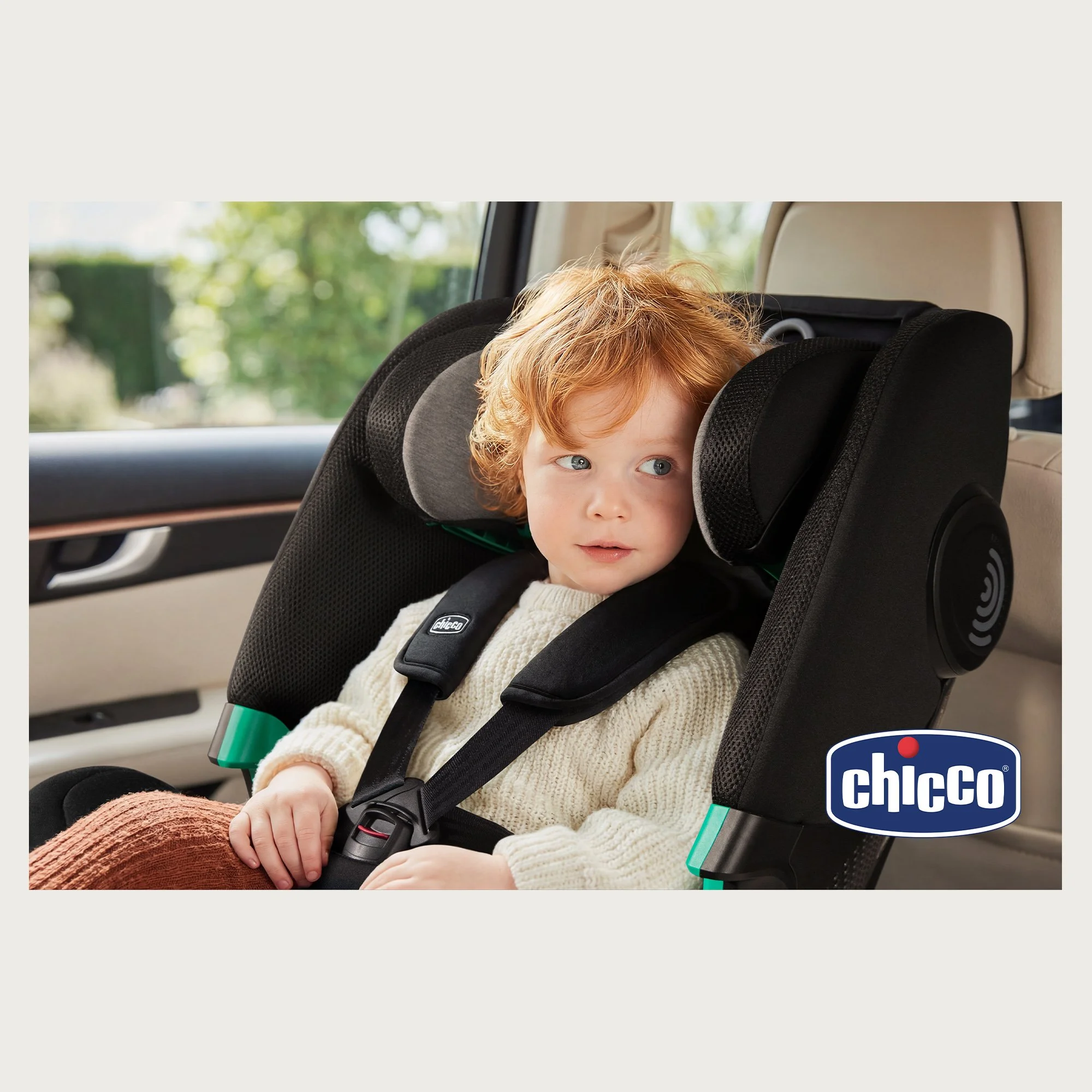 red-haired toddler strapped into a black car seat, natural light. Chicco product.