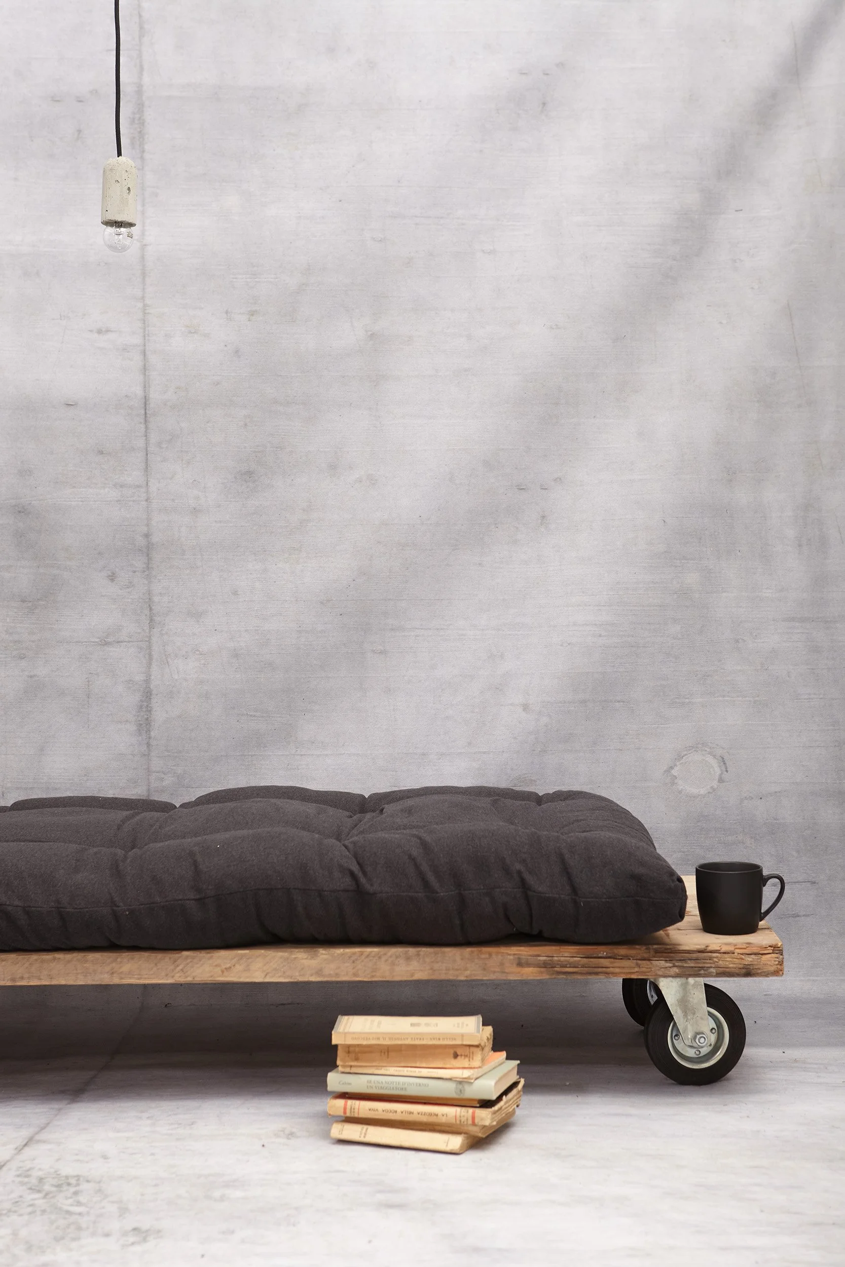 Wooden daybed with dark futon, stacked books and mug against a concrete wall