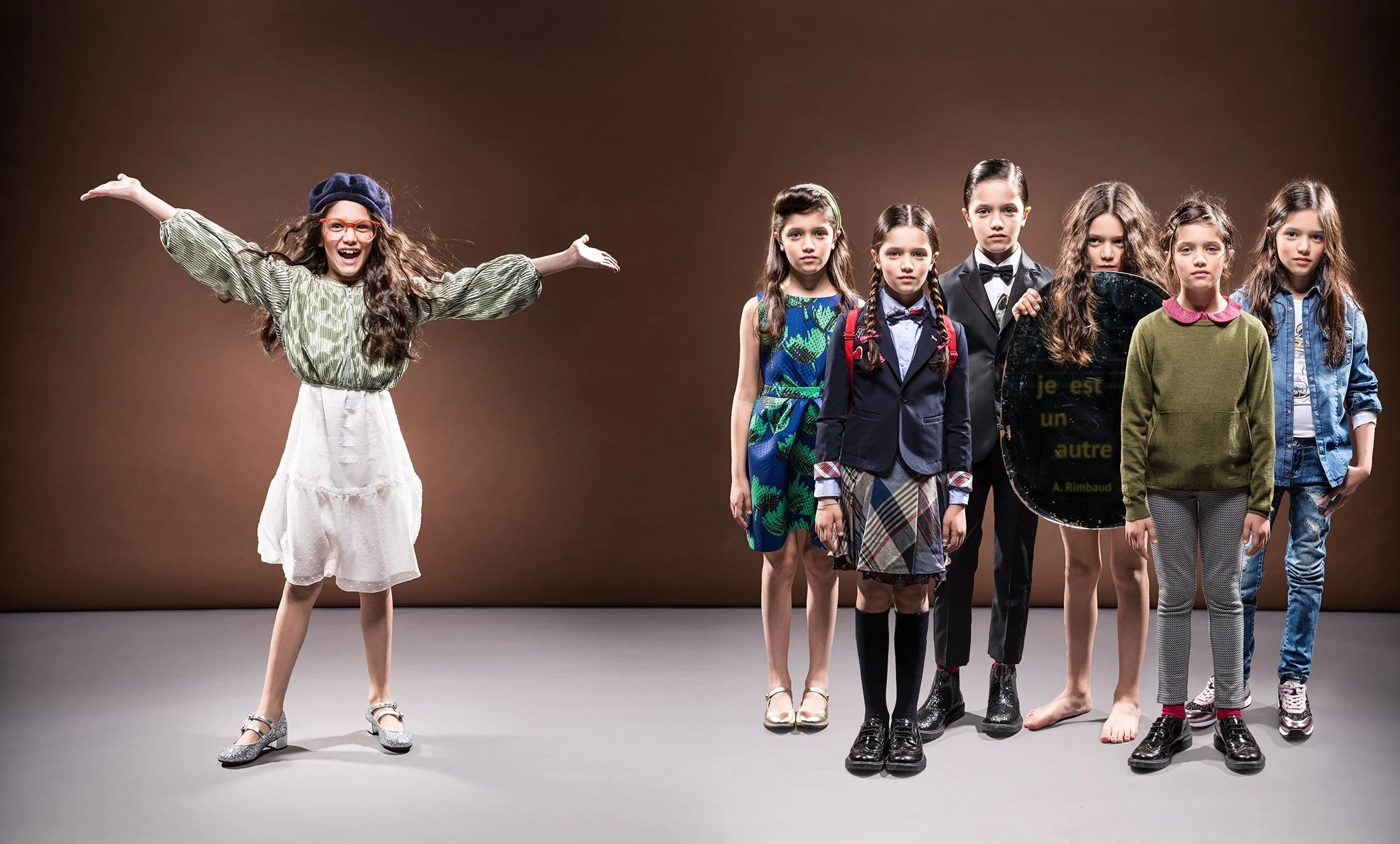One girl in different looks against a brown seamless, from tailoring and schoolwear to knitwear and denim, with a mirror reading “je est un autre” — kidswear editorial, composite studio portrait.