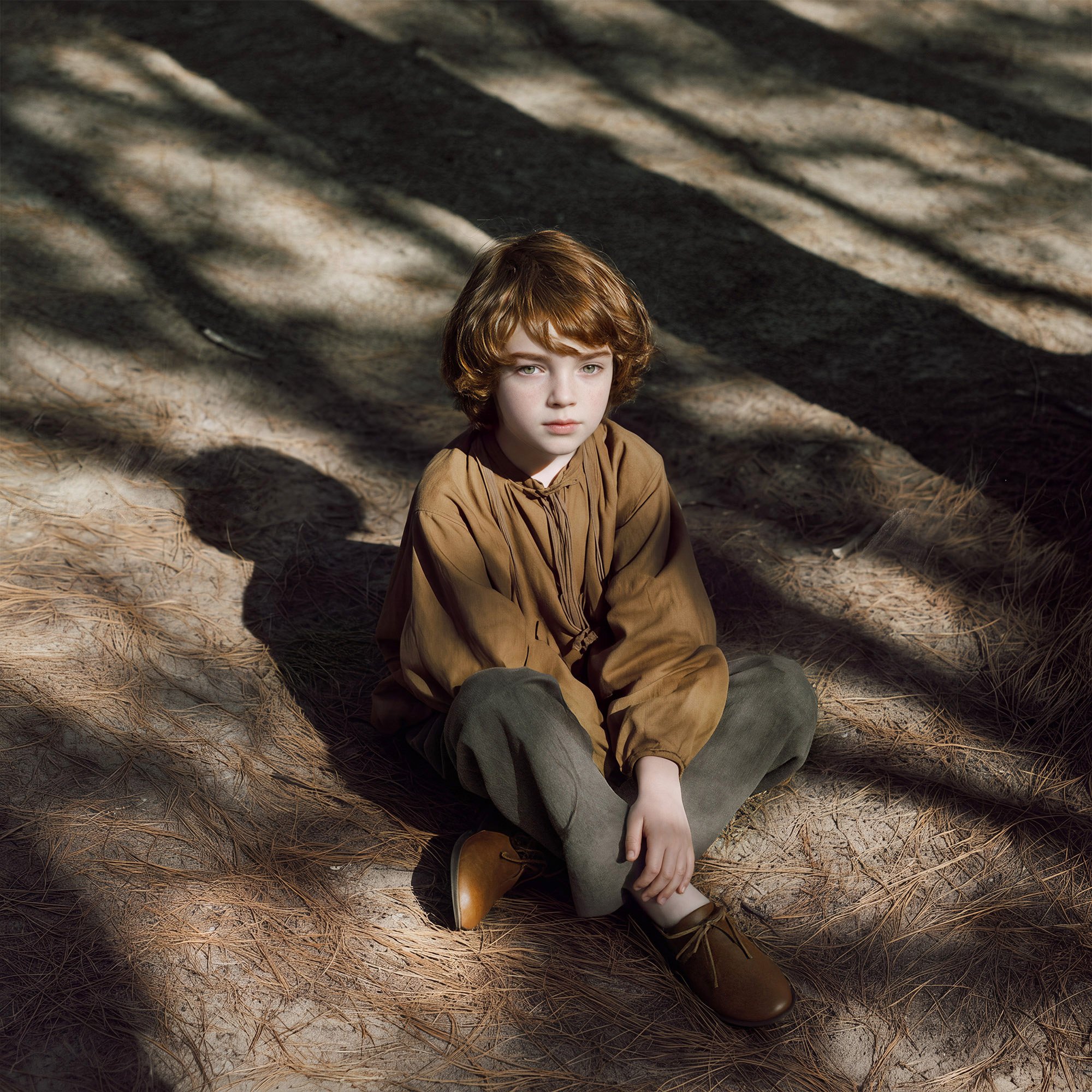 Child sitting cross-legged on pine needles in a mustard linen smock and grey trousers, long tree shadows across the ground — kidswear editorial, warm earth palette, AI-generated image.