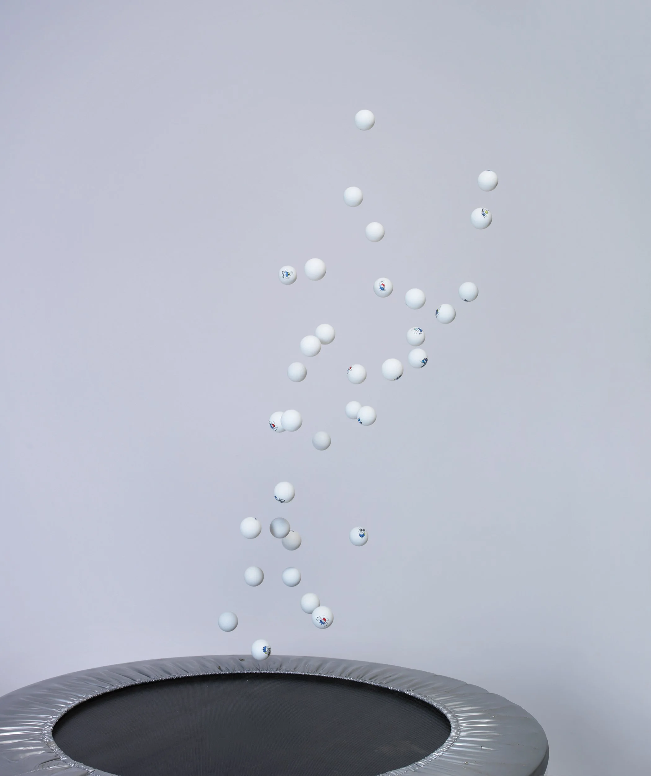 Ping-pong balls suspended in mid-air above a black trampoline bed on a pale grey ground — constructed still life, movement and scatter, studio.