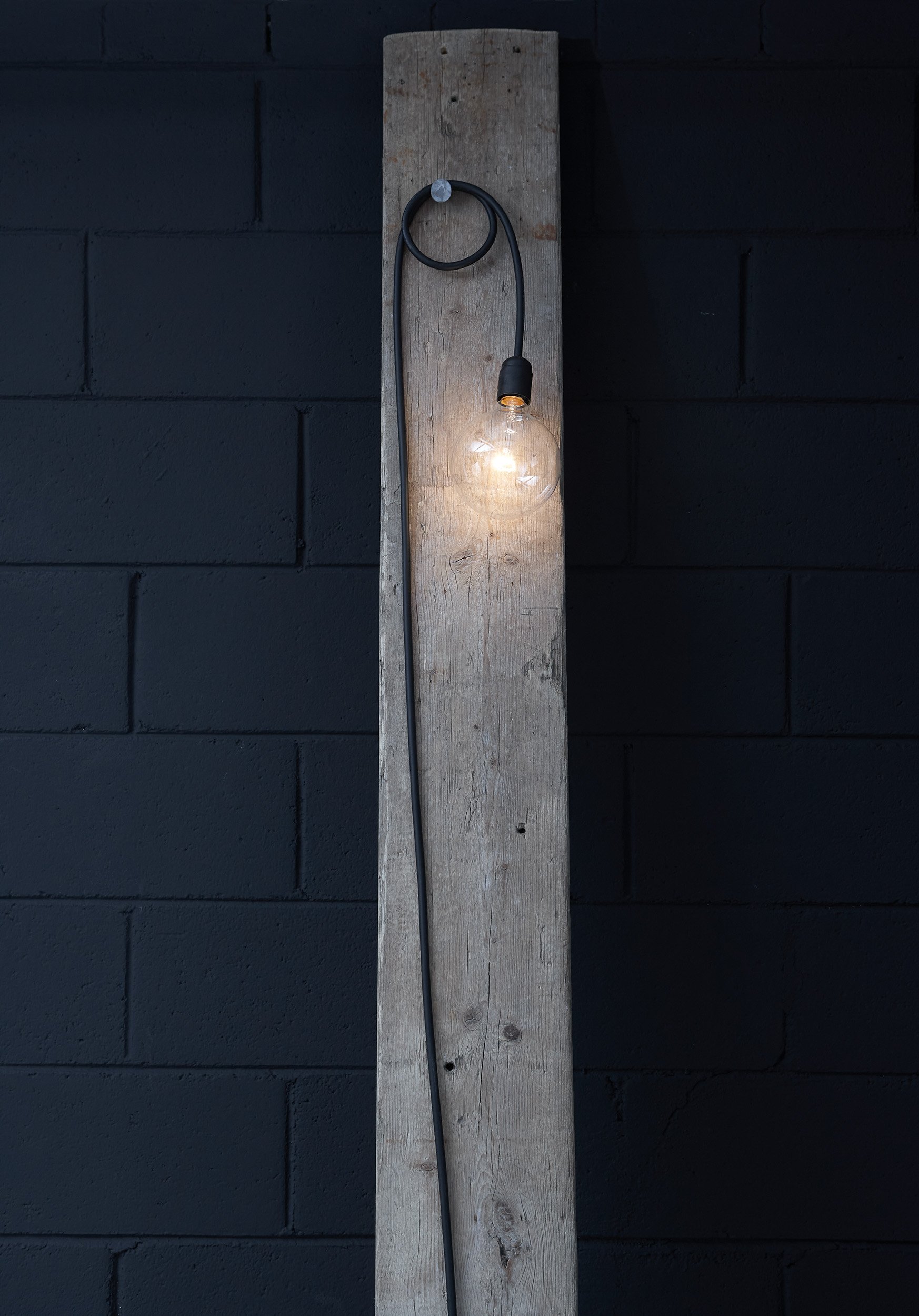Bare globe bulb on a black cable looped over a nail, hung against a weathered reclaimed wood beam and a dark painted breeze-block wall — interiors, lighting detail.