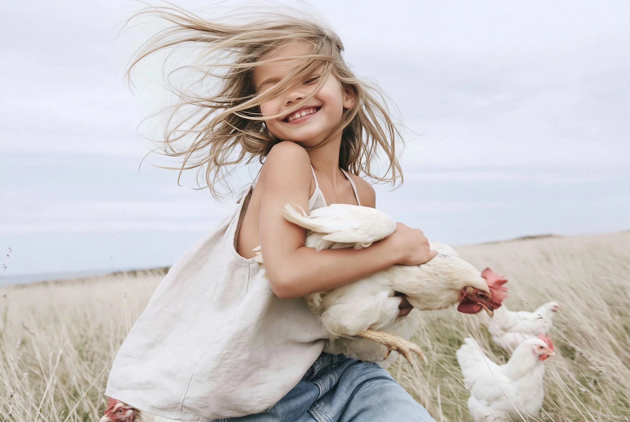 Girl running through dry grass holding a hen, linen cami top and denim shorts, hair in the wind — kidswear lifestyle campaign, overcast daylight, AI-generated image.