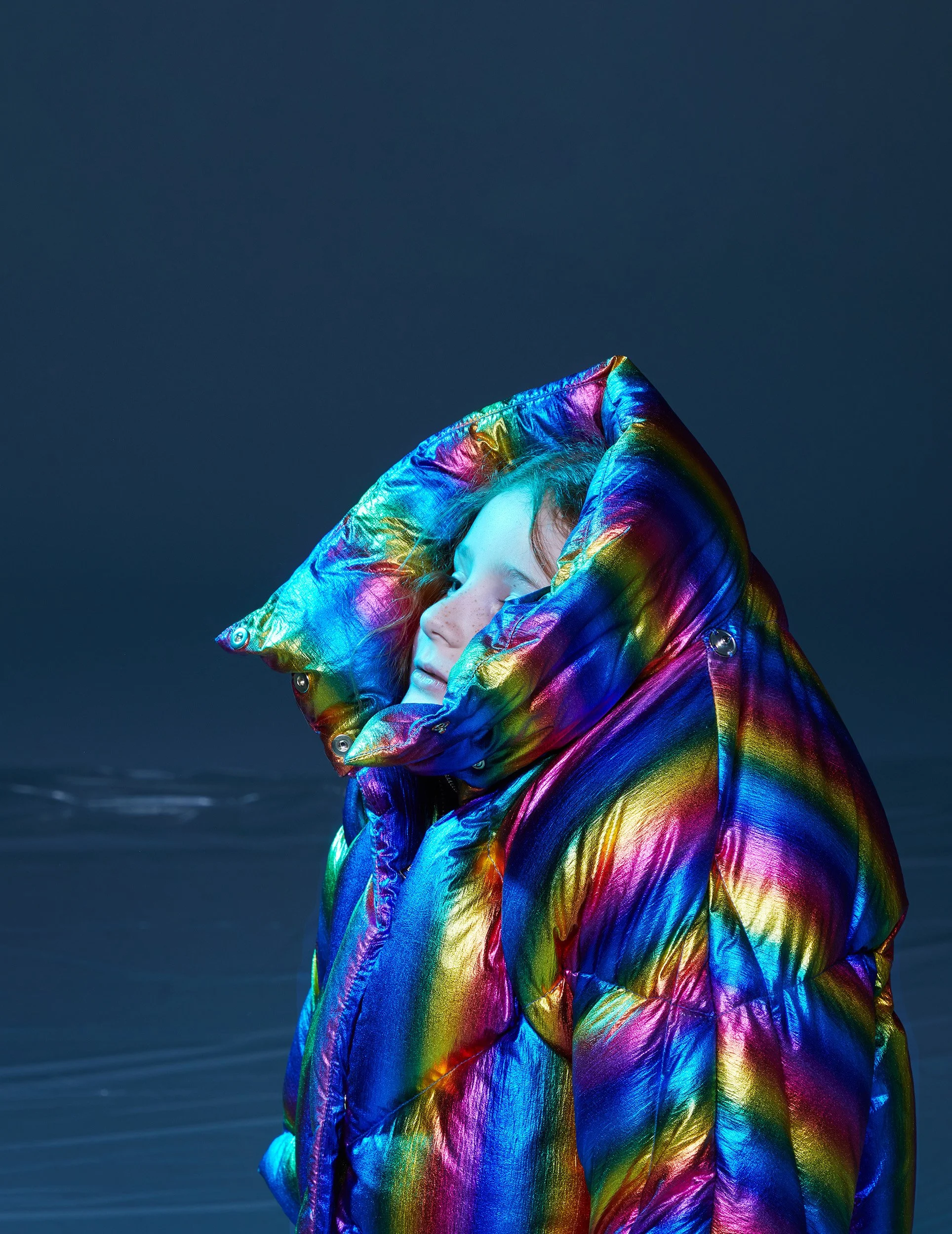 Child in profile inside an oversized hooded puffer in iridescent rainbow nylon, eyes closed against a deep teal ground — kidswear editorial, metallic fabric, studio.