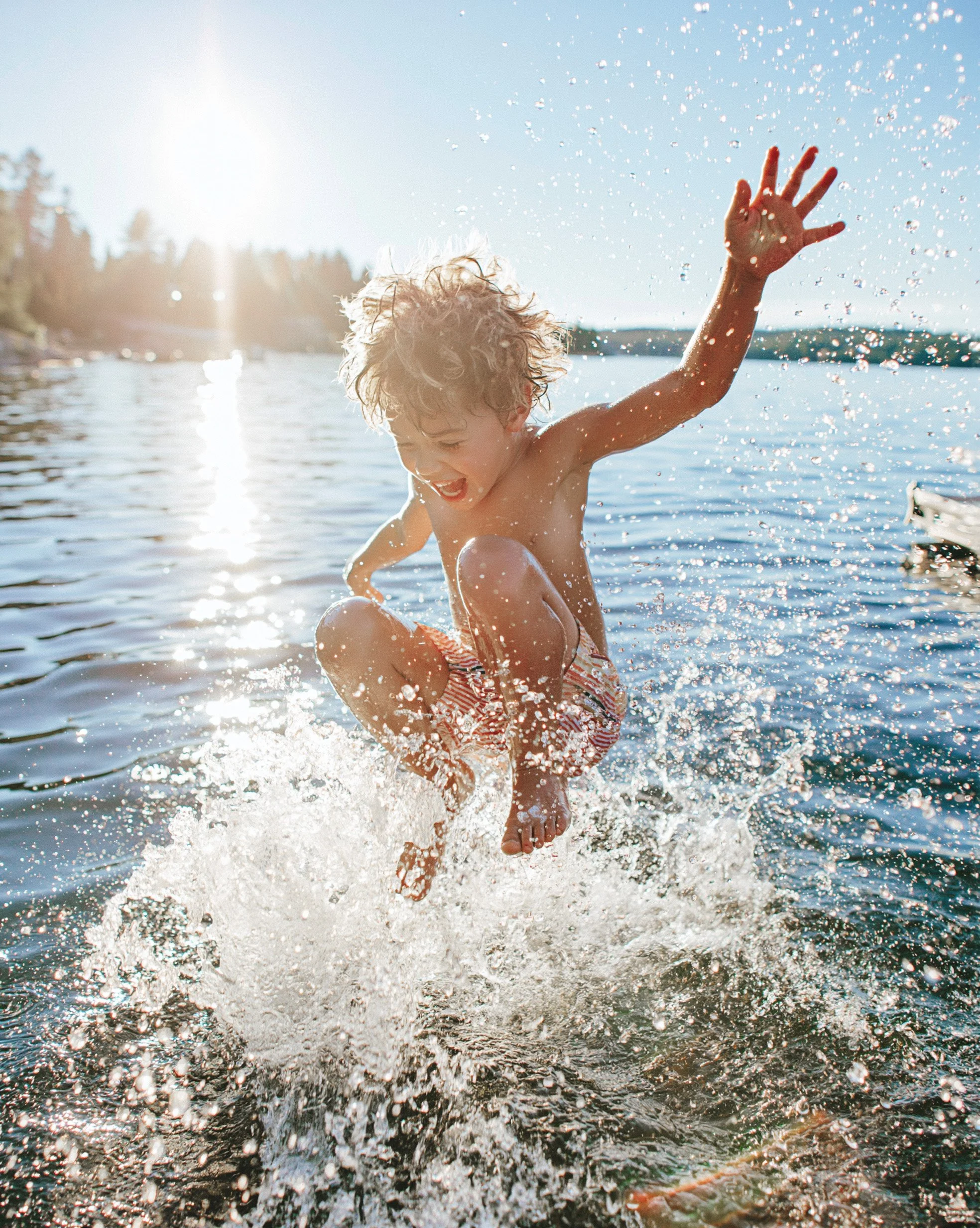 Child leaping into a lake in striped swim shorts, splash and backlight over a pine shoreline — kidswear swimwear campaign, summer, AI-generated image.