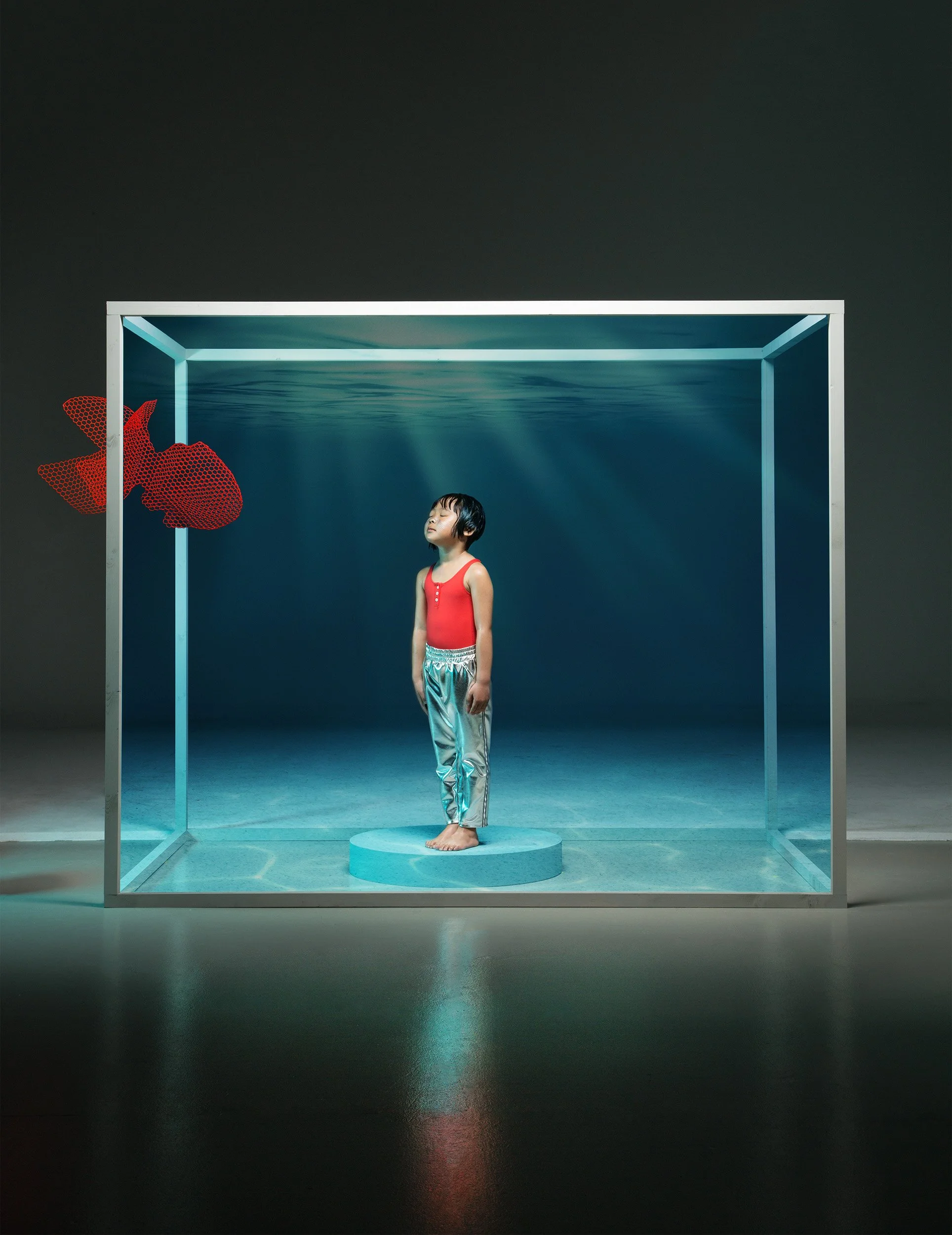 Style Piccoli editorial — a girl inside a glass tank, red fish suspended in the dark.