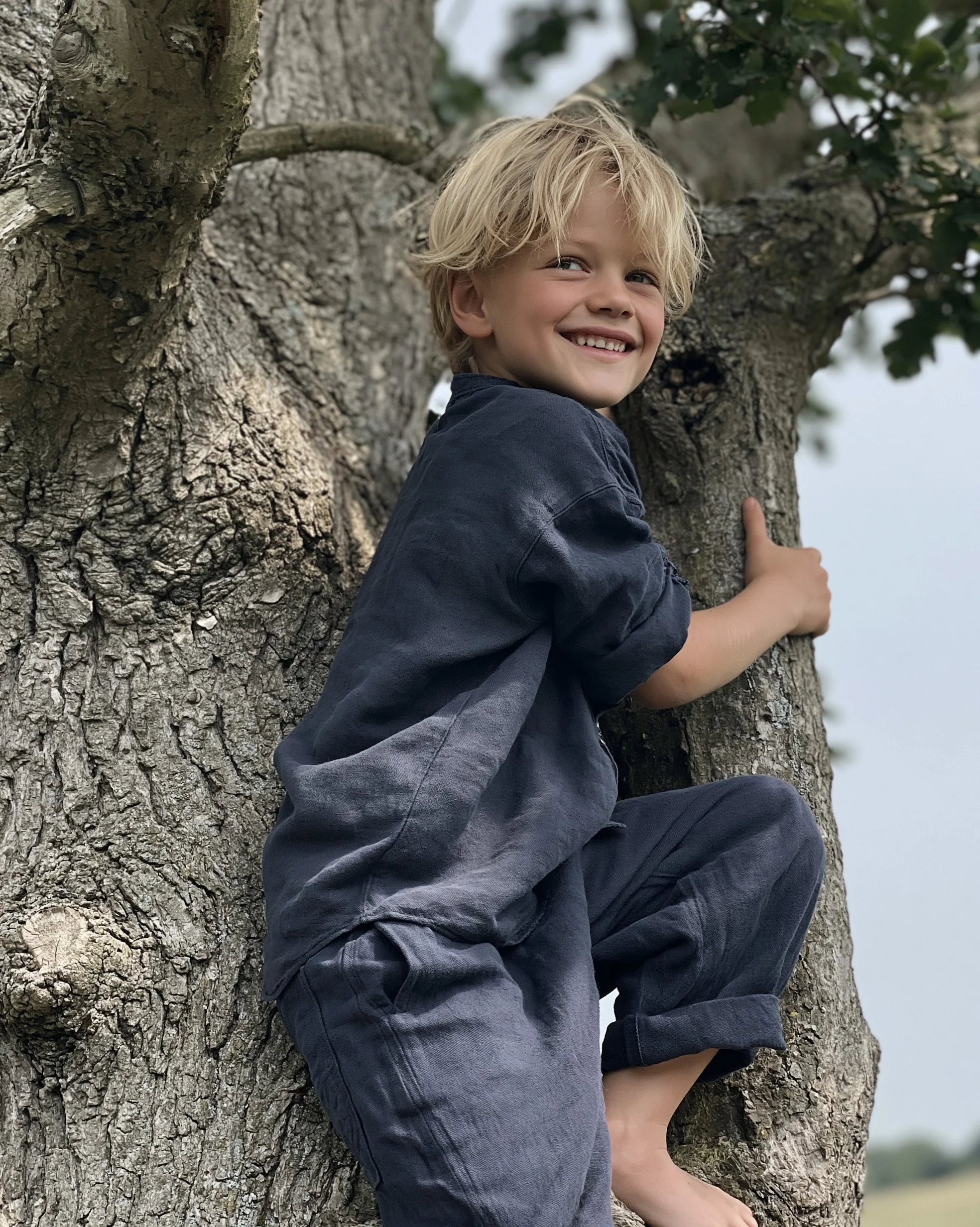 Boy in dark blue linen climbing an old tree, barefoot. AI-generated image.