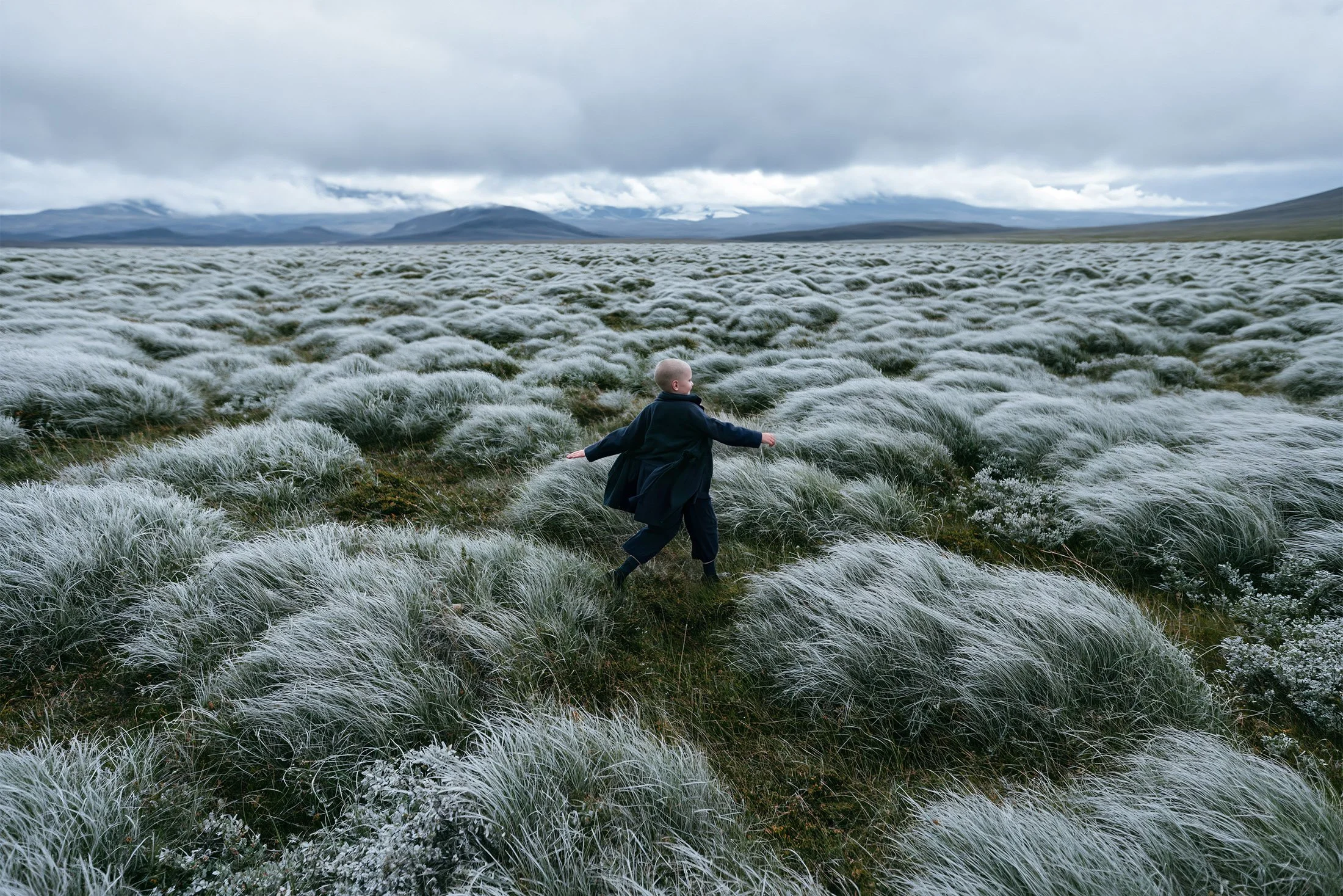 Child in a navy coat and trousers walking arms out through an Icelandic moss field under low cloud — kidswear outerwear campaign, wide landscape, AI-generated image.