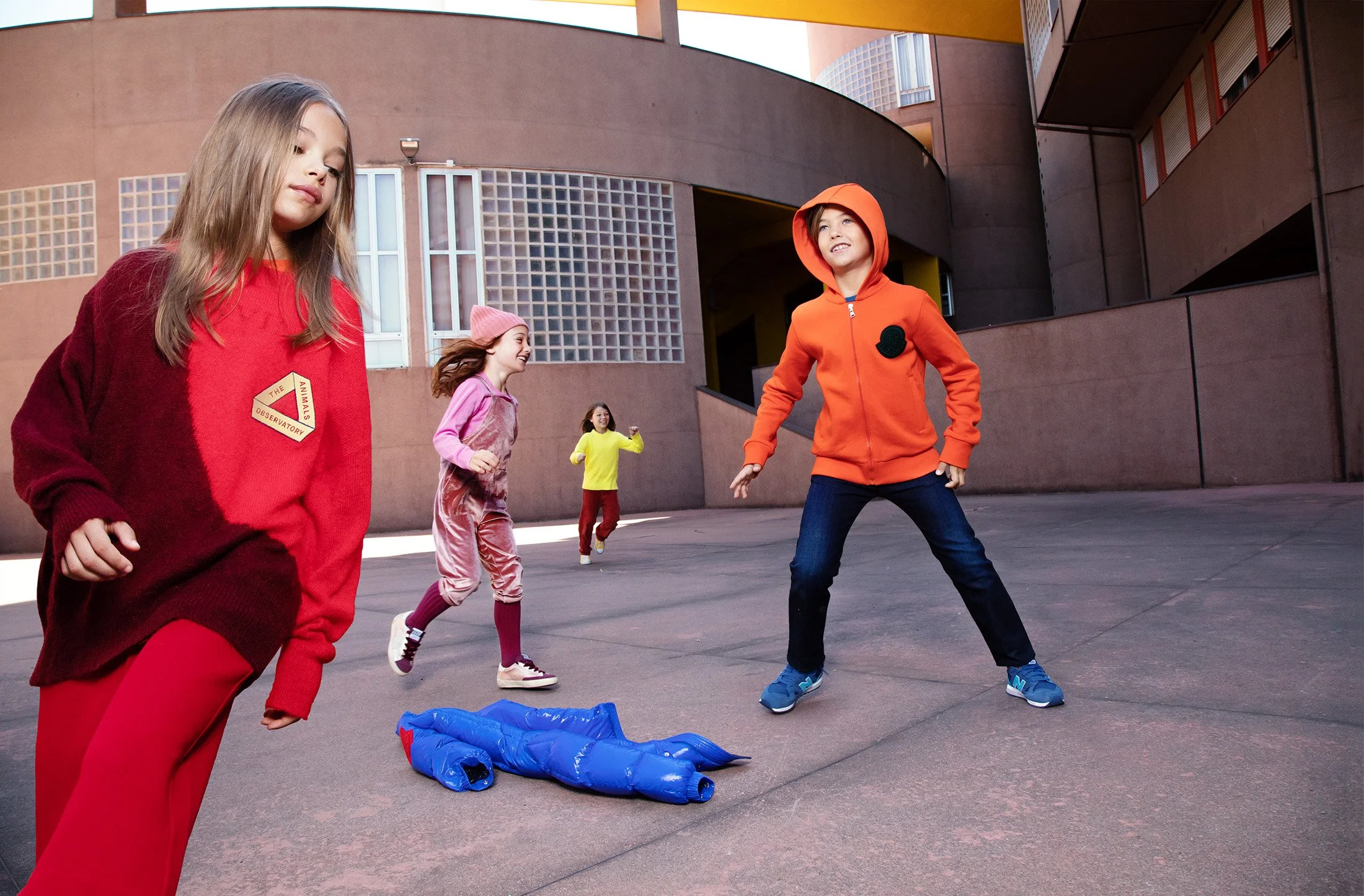 Style Piccoli editorial — children playing in red sportswear in a brutalist courtyard.