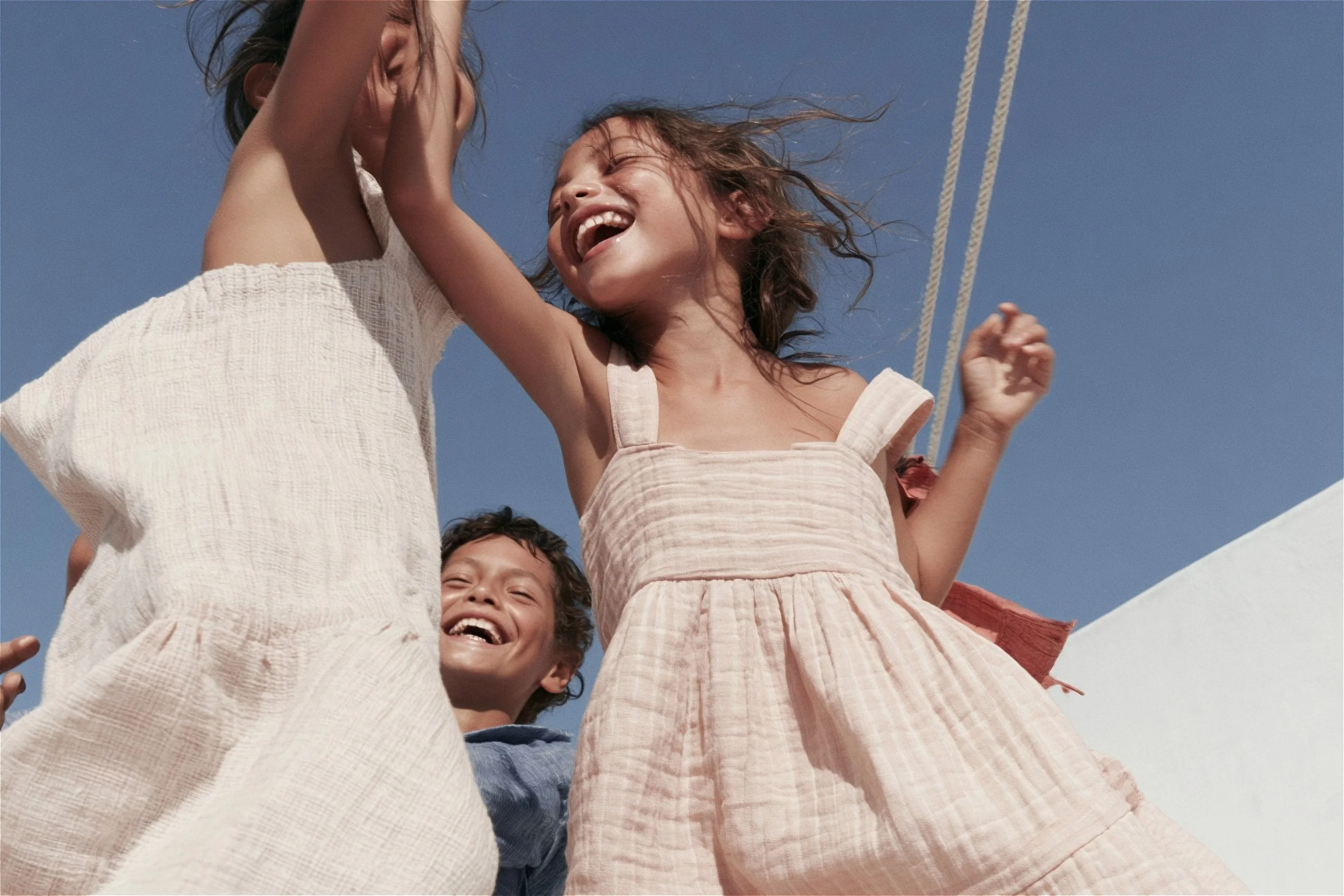 Children laughing on a swing against a deep blue sky, crinkle cotton sundresses in cream and pink — kidswear summer campaign, low angle, AI-generated image.