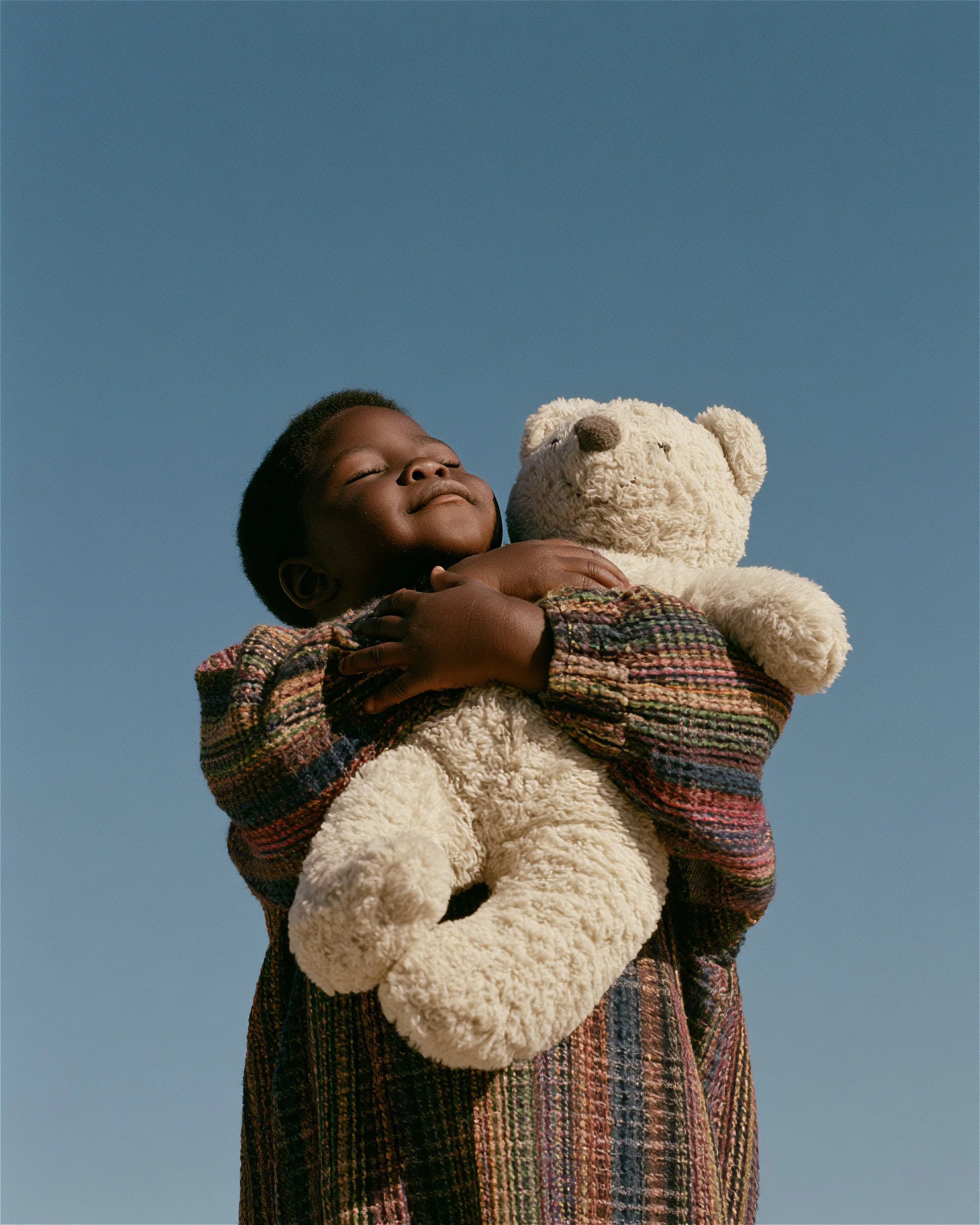 Small girl in a check coat holding a teddy bear against open blue sky. AI-generated image.