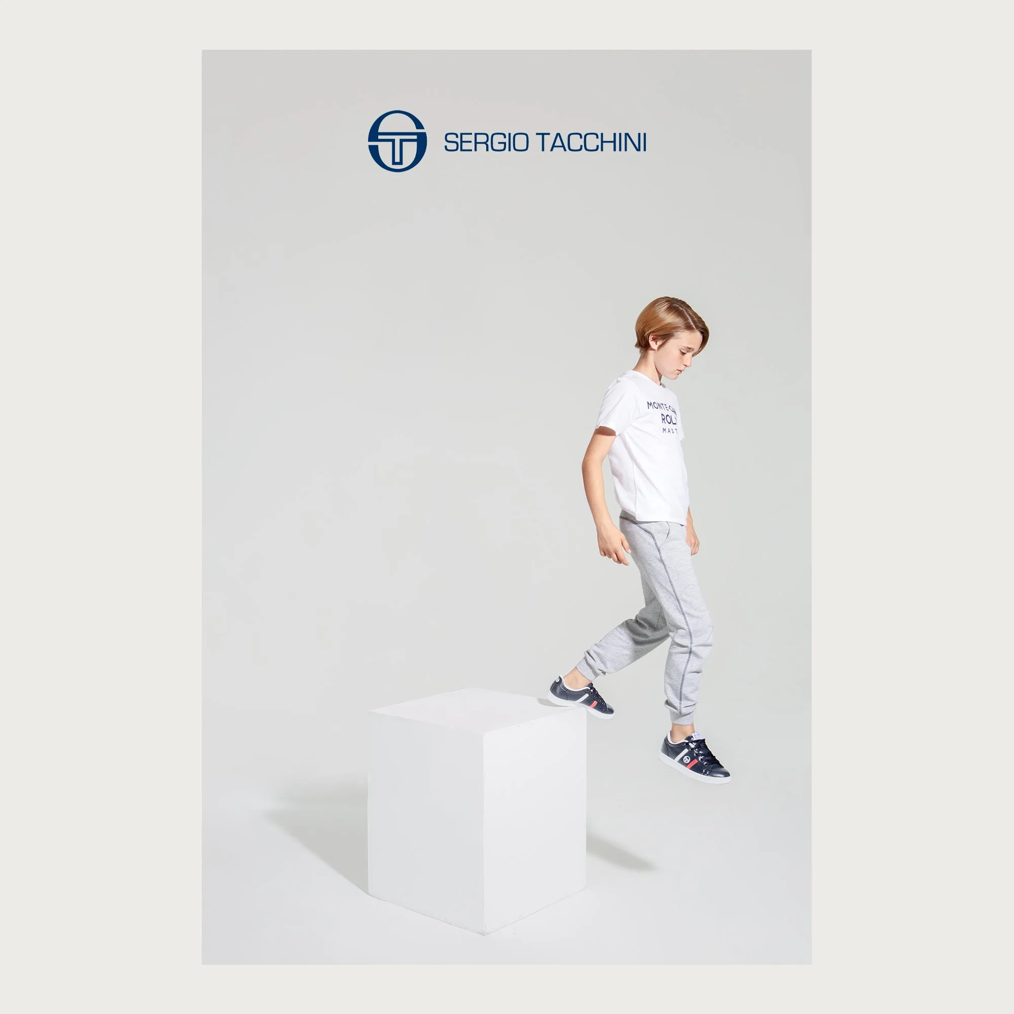 Navy leather sneakers and grey joggers, boy stepping off a white block — Sergio Tacchini kids campaign.