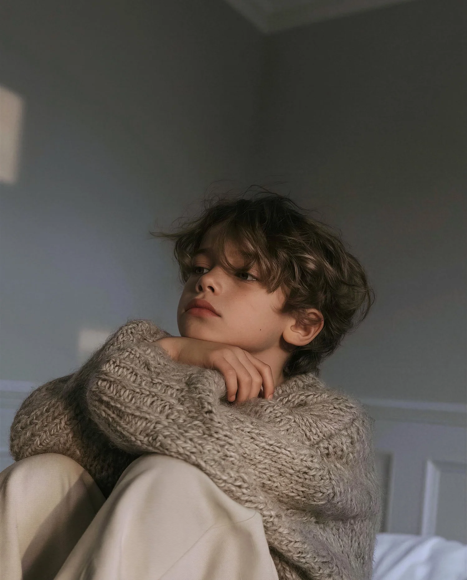 Child in a chunky hand-knit wool sweater and cream trousers, pale blue panelled room — kidswear knitwear editorial, soft window light, AI-generated image.