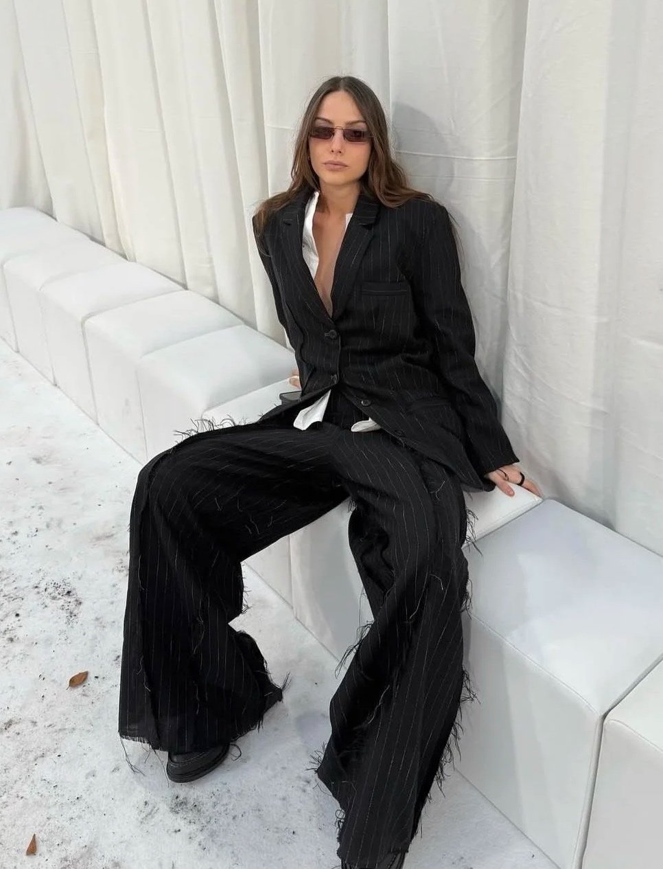 A woman with long hair wearing a black pinstripe suit, sunglasses, and black shoes, sitting on a white bench against white curtains, with a neutral expression.