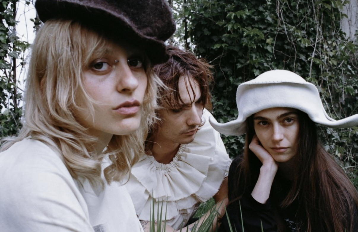 Three women outdoors, one with blonde hair wearing a dark beret, another with reddish hair and ruffled white blouse, and the third with long dark hair and a large white hat, surrounded by green foliage.