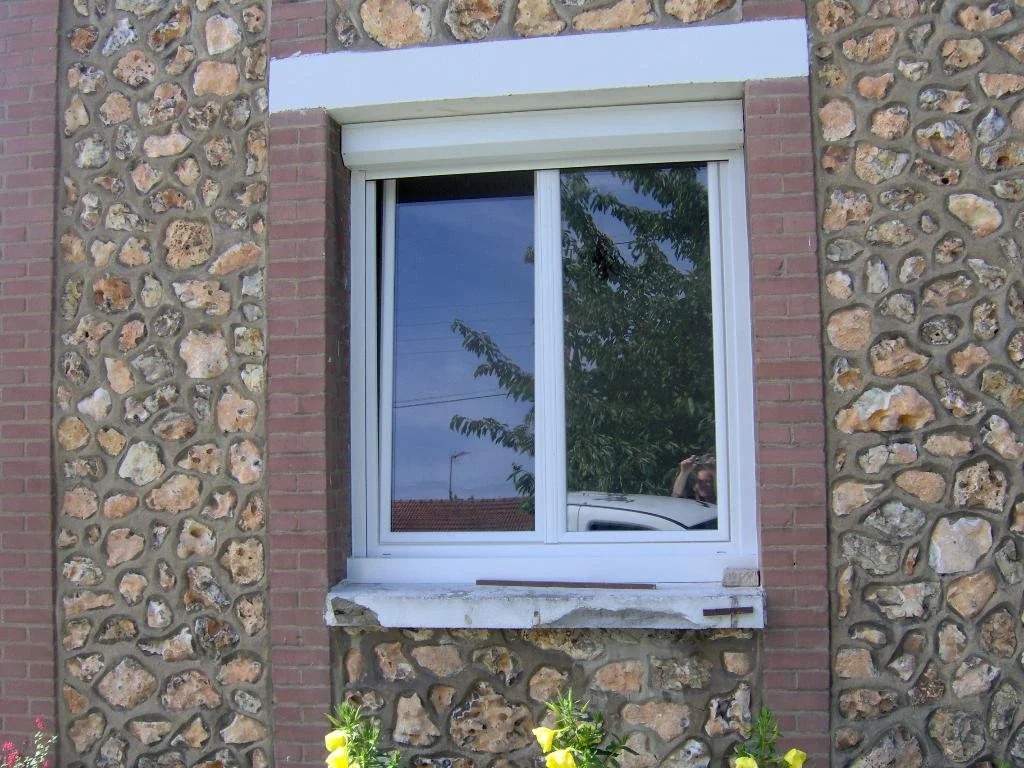 Fenêtre blanche à double vantail avec volet roulant, encadrée de murs en pierre et brique, avec reflection d'arbres et de voitures.