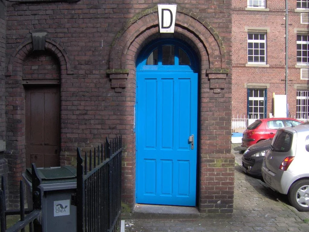 Une porte bleue en bois dans un mur en briques rouges, avec un panneau 'D' au-dessus, située dans une zone urbaine avec des voitures garées et un petit mur avec une barrière noire.