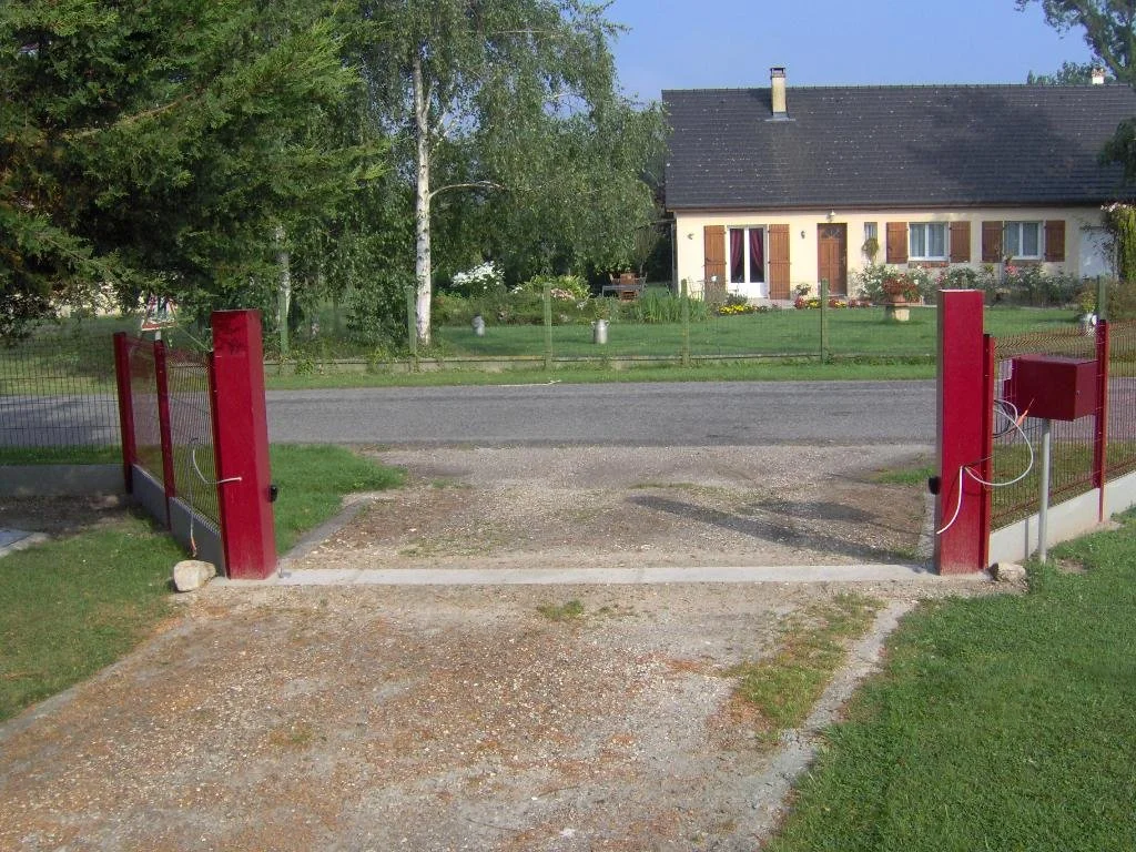 Une petite entrée de jardin avec une clôture rouge et un portail, donnant sur une allée en gravier et une maison avec des volets en bois.