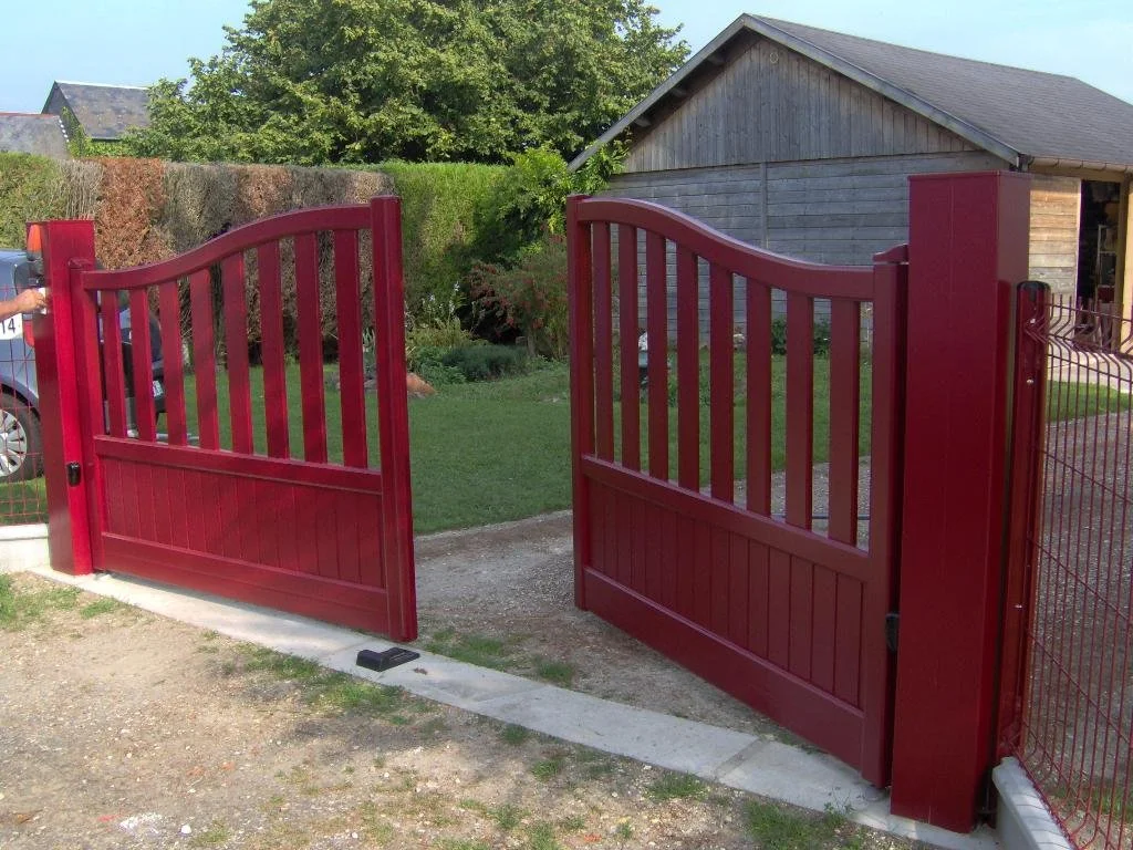 Une porte de clôture en bois peinte en rouge, avec un panneau de clôture en fil métallique rouge à droite, menant à un jardin avec une maison en bois en arrière-plan.