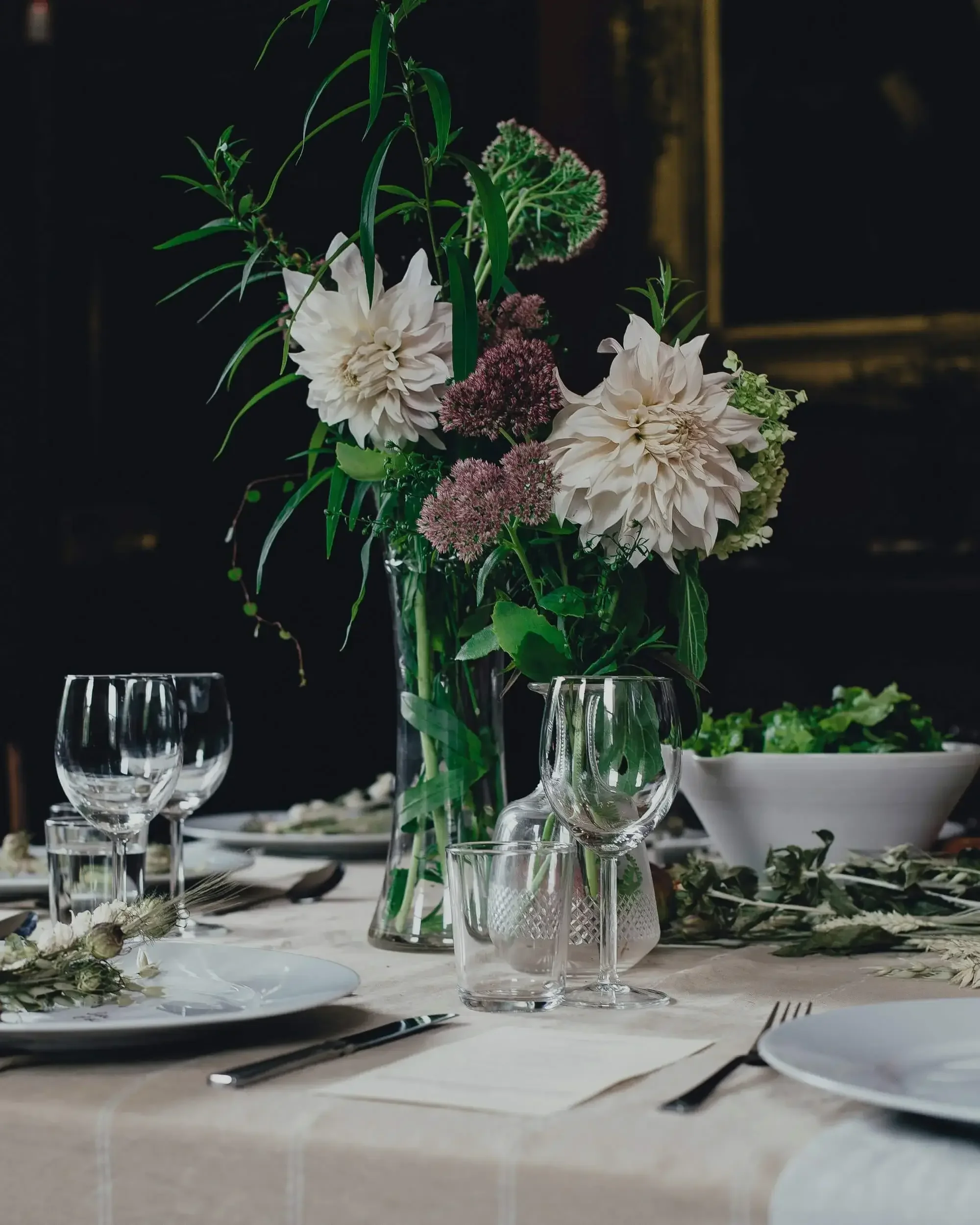 Elegant table setting with tall floral centerpiece containing white and purple flowers, and a mix of greenery in glass vases. Surrounding the centerpiece are empty wine and water glasses, plates, cutlery. Lous Kitchen by Louise Field.