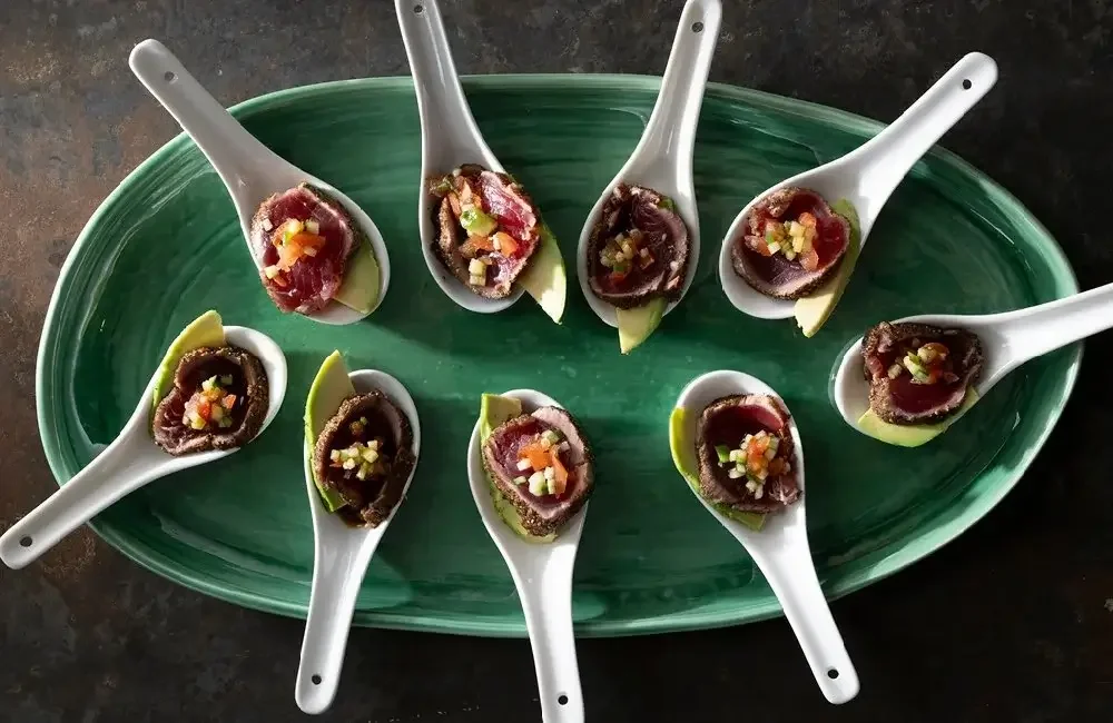 A green oval platter with several white serving spoons, each holding slices of ahi tuna sashimi topped with diced vegetables, and a small wedge of cucumber.