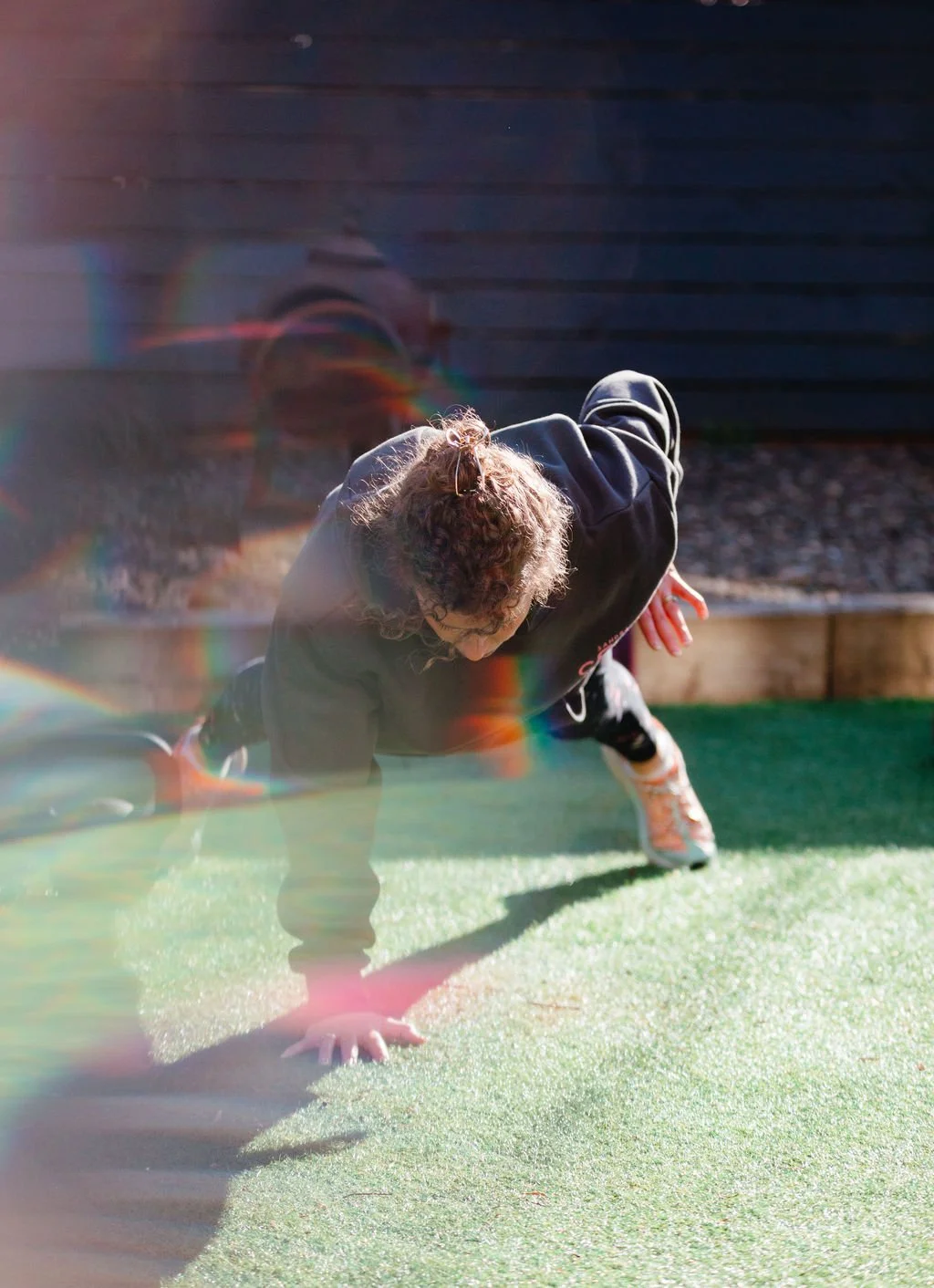 A person with curly hair doing a one-arm push-up outdoors on a grassy area, with a wooden fence in the background and sunlight creating a lens flare.
