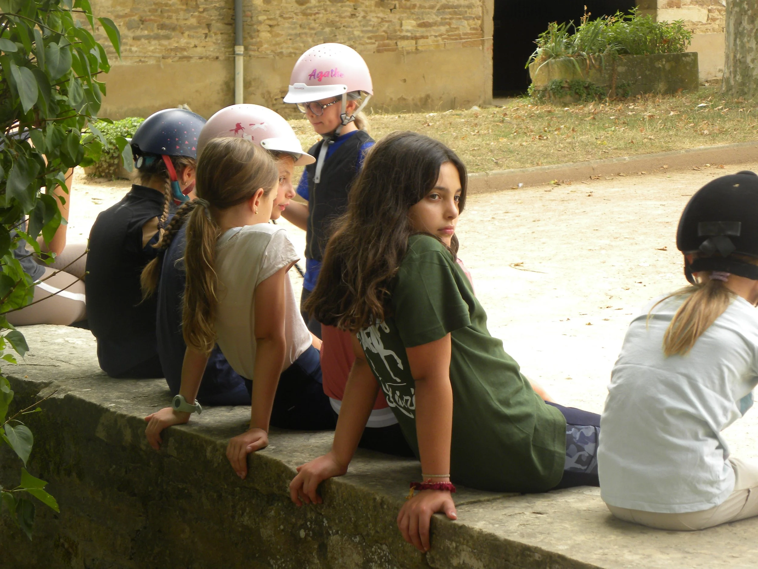 Groupe d'enfants assis en ligne, portant des casques, à l'extérieur près d'un mur en pierre, certains regardant vers l'avant ou vers le bas.