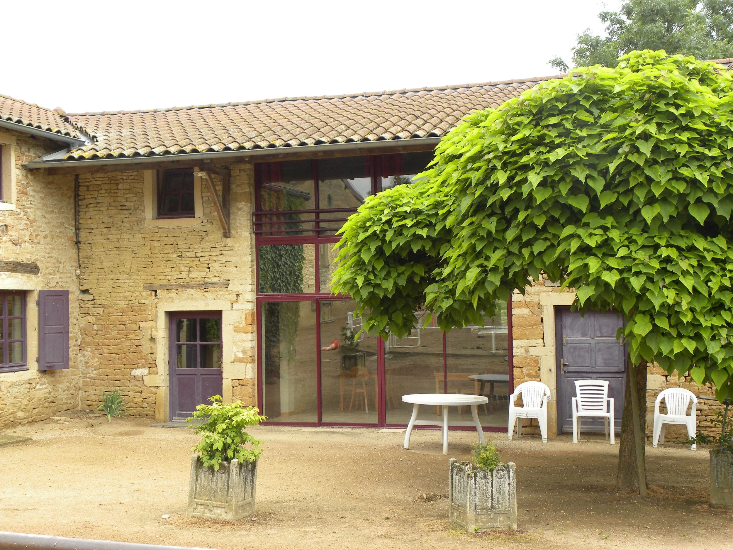 Une maison en pierre avec un toit en tuiles, un balcon en verre avec une structuration en métal rouge, et un grand arbre à feuilles vertes devant. Il y a également des chaises en plastique blanc et une table en métal dans la cour.