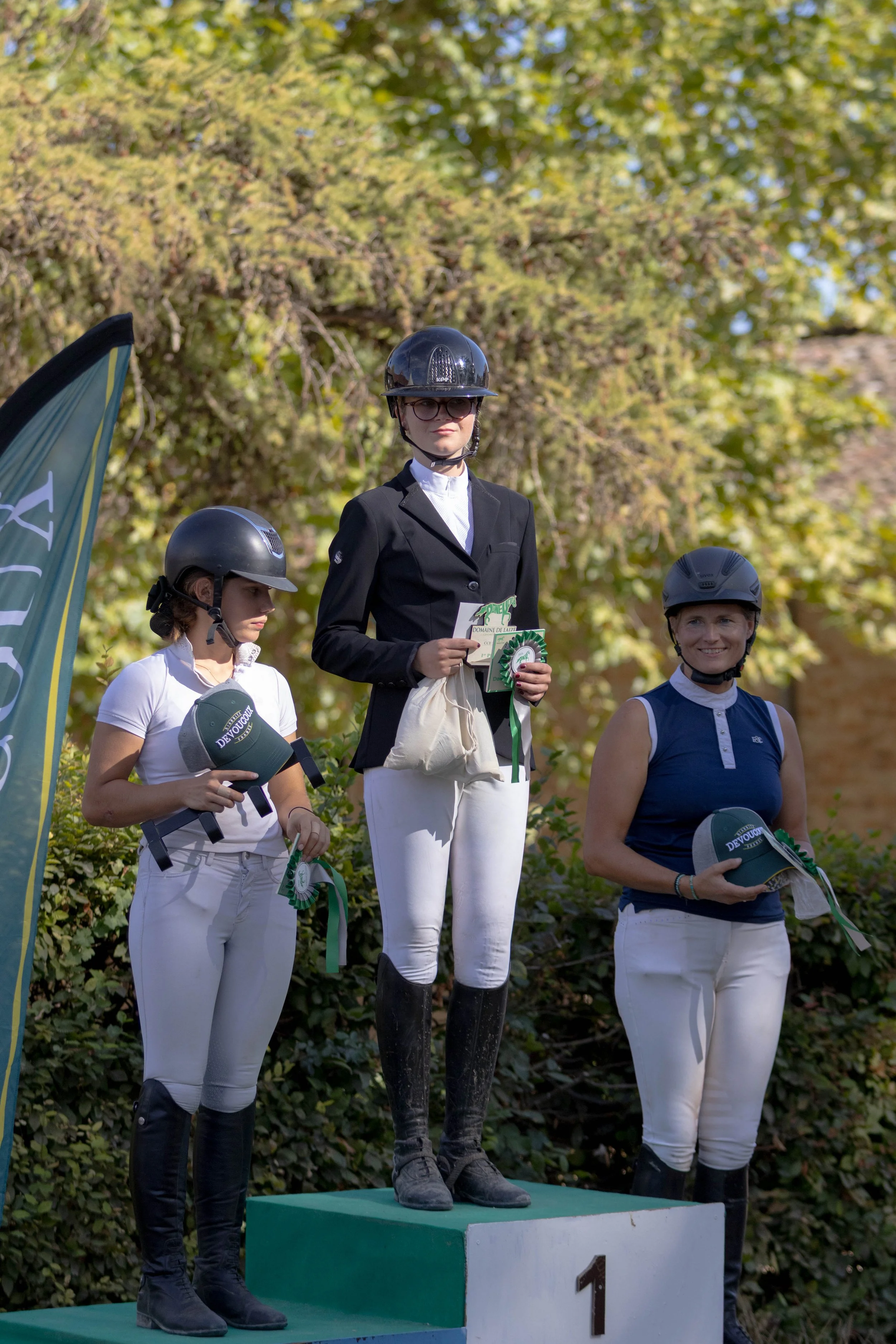 Trois femmes en tenue d'équitation, portant des casques, se tiennent sur un podium de remise de prix en extérieur, avec un arbre en arrière-plan. La femme au centre est en première position, tenant une carte et une rosette verte, et porte une veste noire et une chemise blanche. La femme à gauche porte une chemise blanche et un casque gris, la femme à droite porte une tenue bleue et blanche.