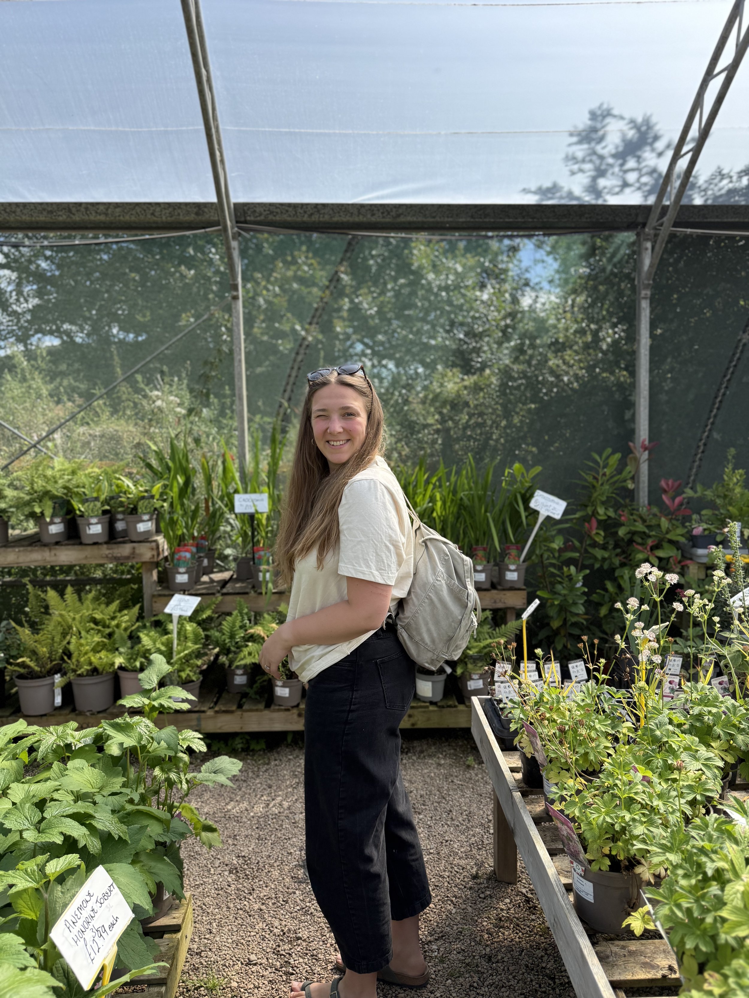 A young woman with long hair, wearing a beige t-shirt, black pants, and sandals, is standing in a plant nursery surrounded by potted plants on wooden tables. She is smiling at the camera under a greenhouse roof with trees visible in the background.