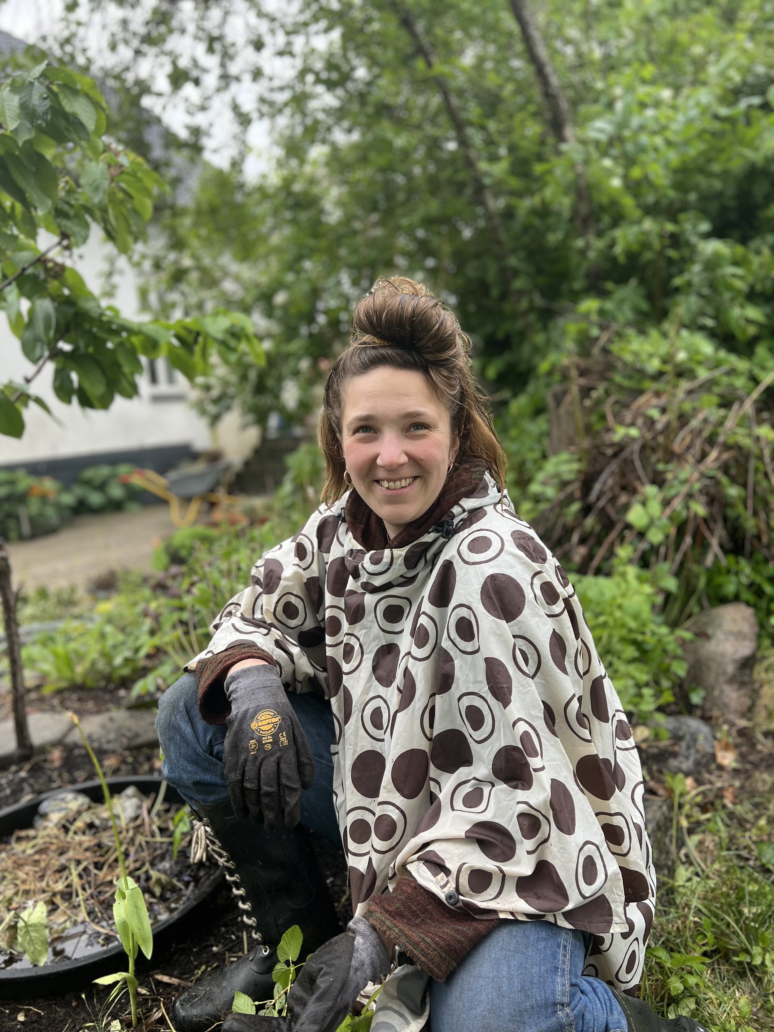 Woman gardening outdoors in a garden with green plants and trees, wearing a polka dot patterned rain cape, gloves, and jeans.