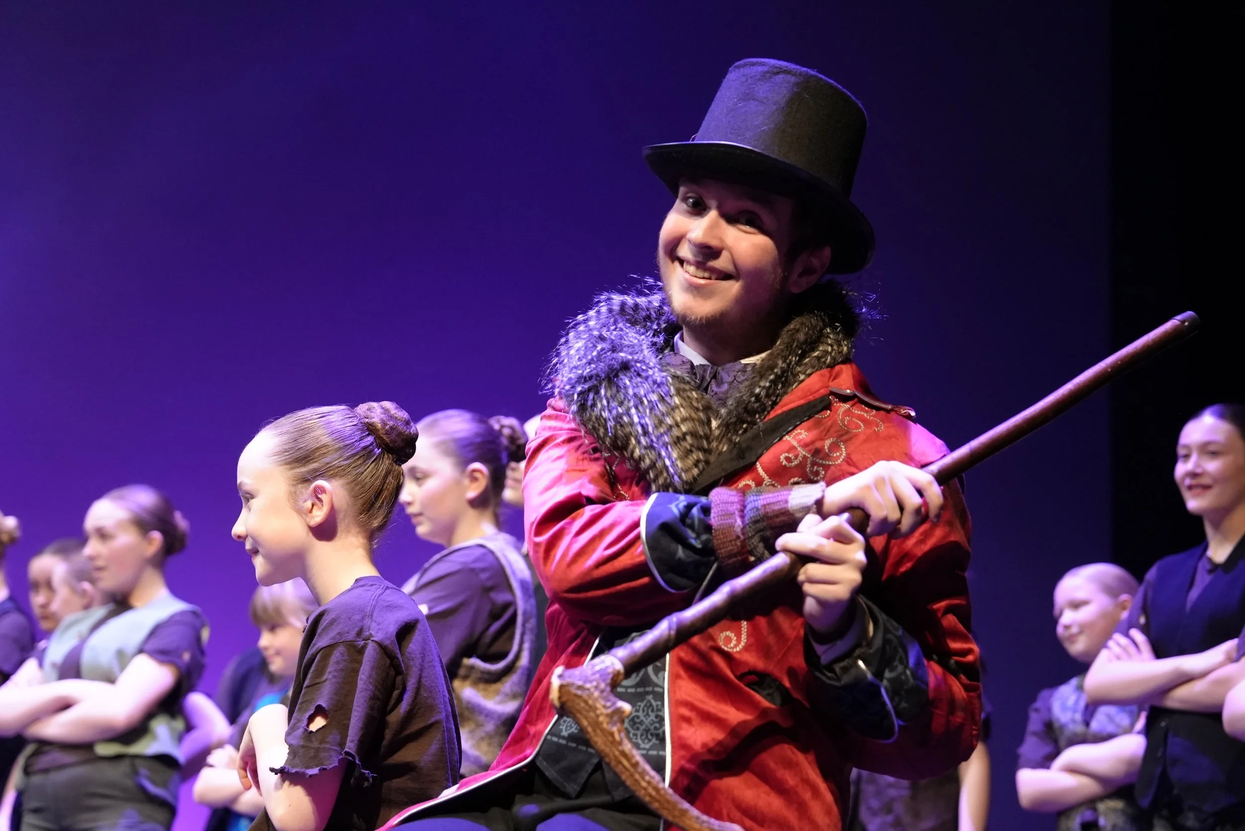 A performer dressed as a ringmaster with a top hat and colorful coat, smiling with children rehearsing or performing in the background.