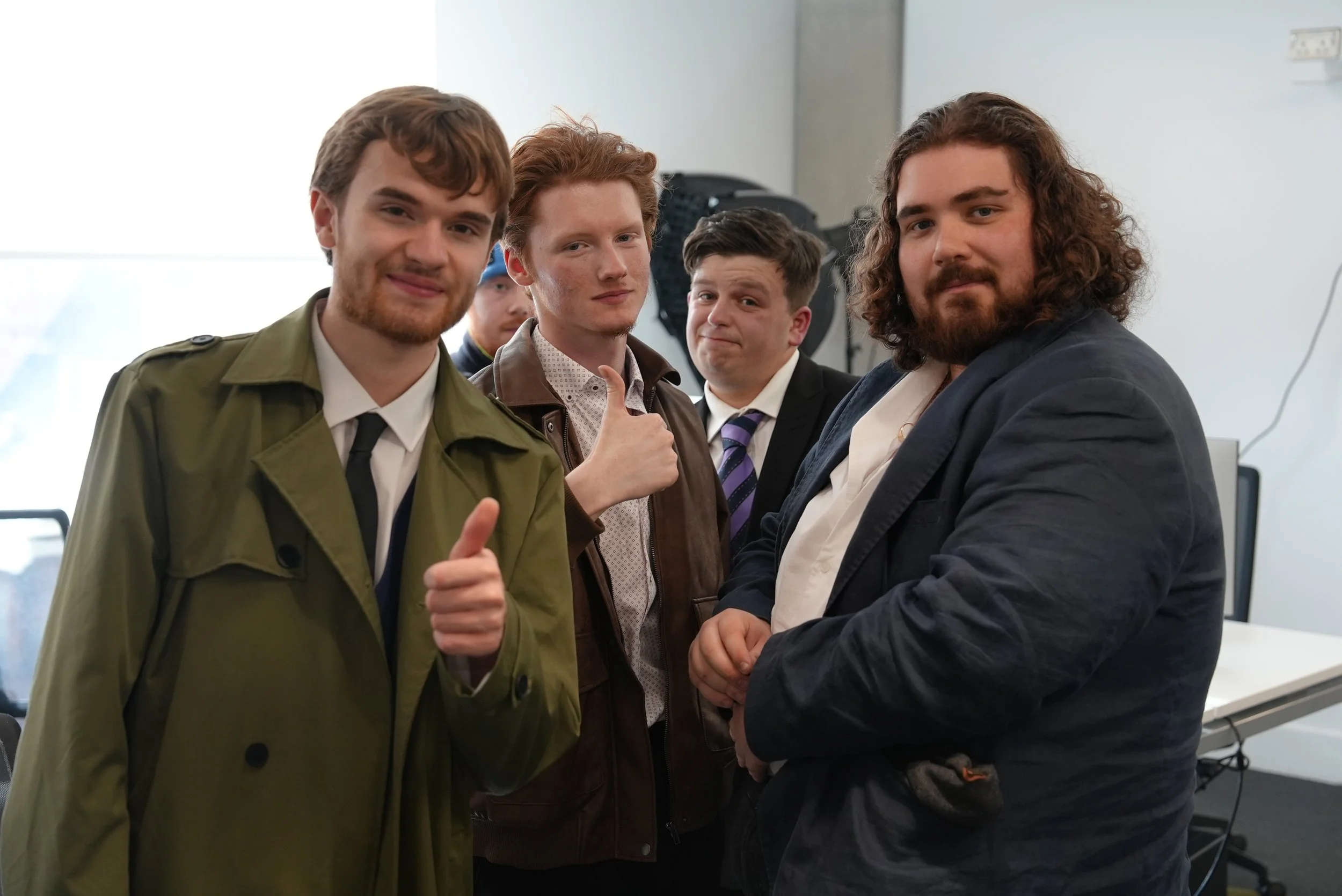 Four young men in business attire standing indoors, some giving thumbs-up, in front of a whiteboard and camera equipment.