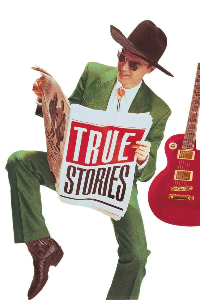A man wearing a green suit, black hat, and sunglasses holding a newspaper with the headline 'TRUE STORIES', with a red guitar standing next to him.