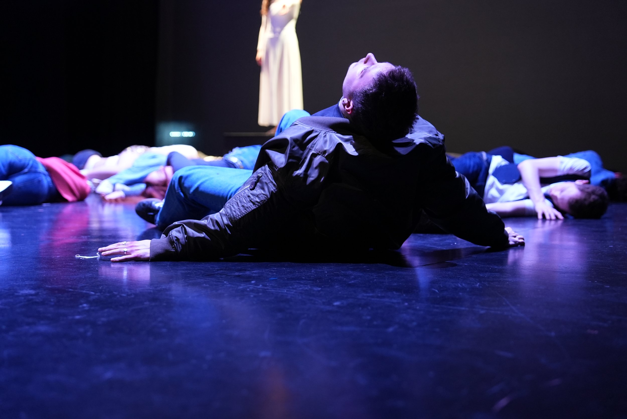 Person in dark clothing lying on the stage floor in a theater, with multiple other people lying on the stage behind them, and a woman standing in the background.